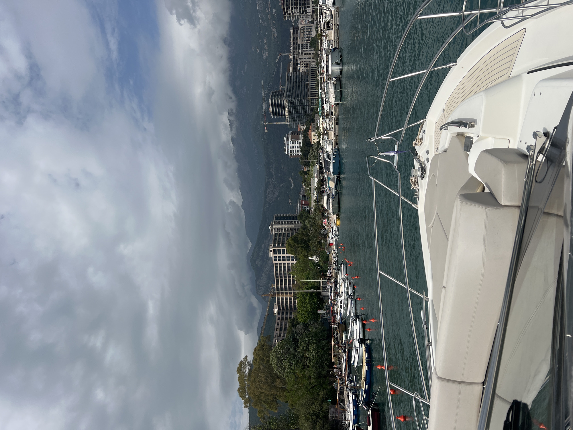 The image depicts a view from a boat docked in a marina along the coast of Budva, Montenegro. Buildings line the shore, including multi-story structures and some under construction. Mountains rise behind the buildings, partially obscured by clouds. Several boats are moored in the marina, delineated by orange buoys. The foreground features the railing, seating, and other parts of the boat from which the photo was taken. The sky is overcast, suggesting a cloudy day.