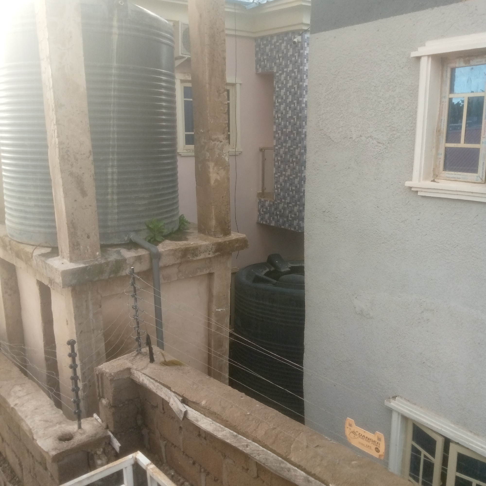 The image displays an exterior scene in the city of Unknown, Unknown, characterized by building infrastructure and utility elements. Two large, corrugated, grey cylindrical water tanks are positioned on a concrete platform in the upper left-midground. A stout, weathered concrete pillar stands between them. A grey pipe extends from the base of the rightmost large tank. Behind these, a light pink building is visible, featuring a white-framed window and an external air conditioning unit. Part of this building's facade is adorned with a grey and white mosaic tile pattern. Further right, a light grey, textured stucco wall dominates the right side of the frame, containing a white-framed window. Below the water tanks and platform, an electric fence system is installed along a low, rough concrete or brick wall, featuring multiple strands of thin wire supported by black insulators. Another smaller, dark green/black ribbed tank, possibly a water tank with an open or damaged lid, is partially visible at ground level between the pink building and the grey wall. On the lower right section of the grey wall, an orange-yellow sign is affixed, partially legible, indicating "DANGER ELECTRIC FENCE" and "LPG". Small green foliage is observed growing on the concrete platform near one of the large tanks. The scene is illuminated by daylight, with some glare visible at the top. No individuals are present.