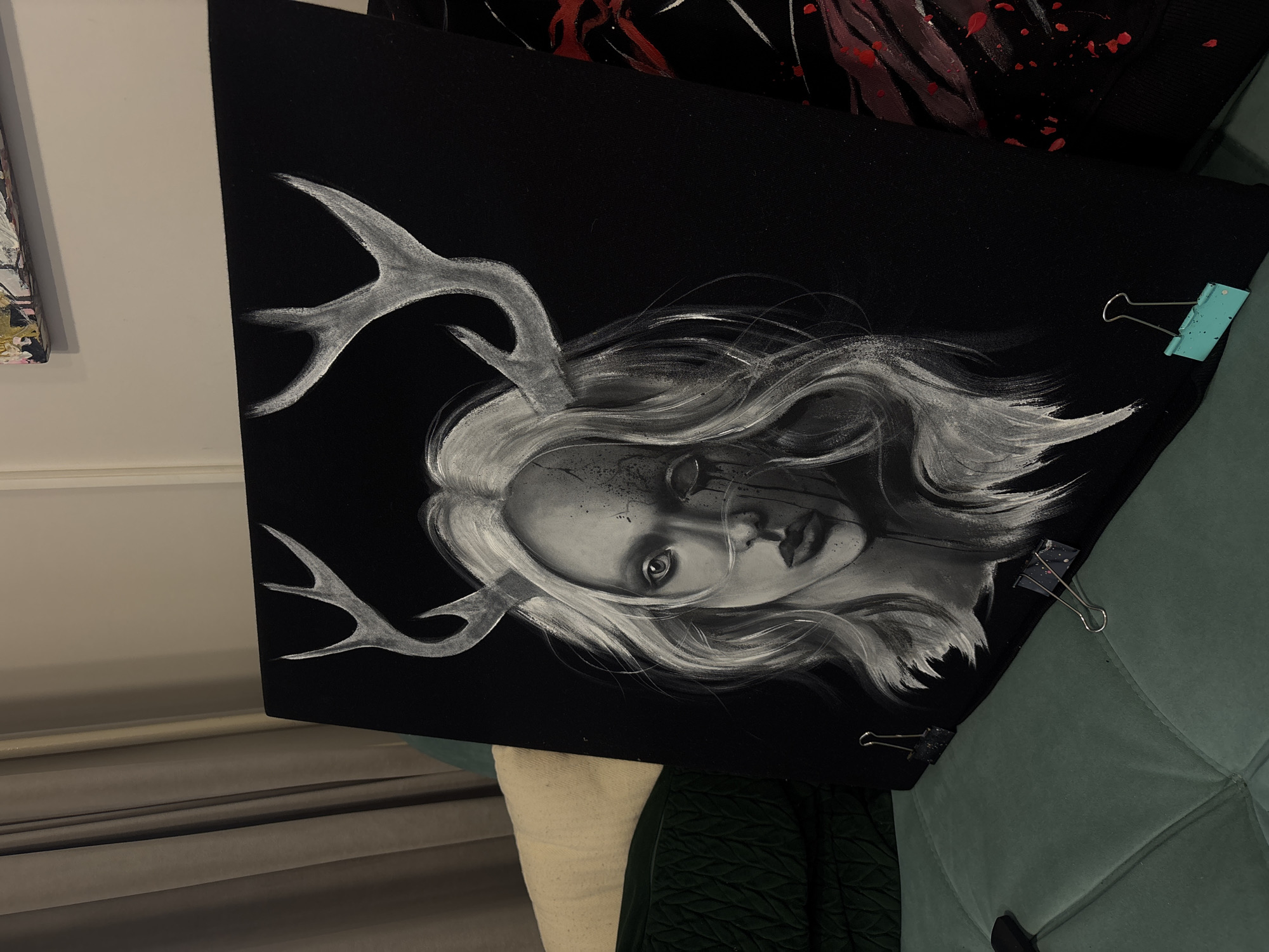 The image shows a painting resting against a green, velvet, tufted surface. The painting is primarily black with a grayscale image of a woman with antlers. The woman has long flowing hair and appears to have cracks on her face with dark liquid running from her eye. The antlers are painted in a similar grayscale tone. The painting is secured with binder clips on the right edge. The background includes a partially visible white wall, a beige blind or curtain, and another painting with red paint splatters. The location is not discernible from the image.