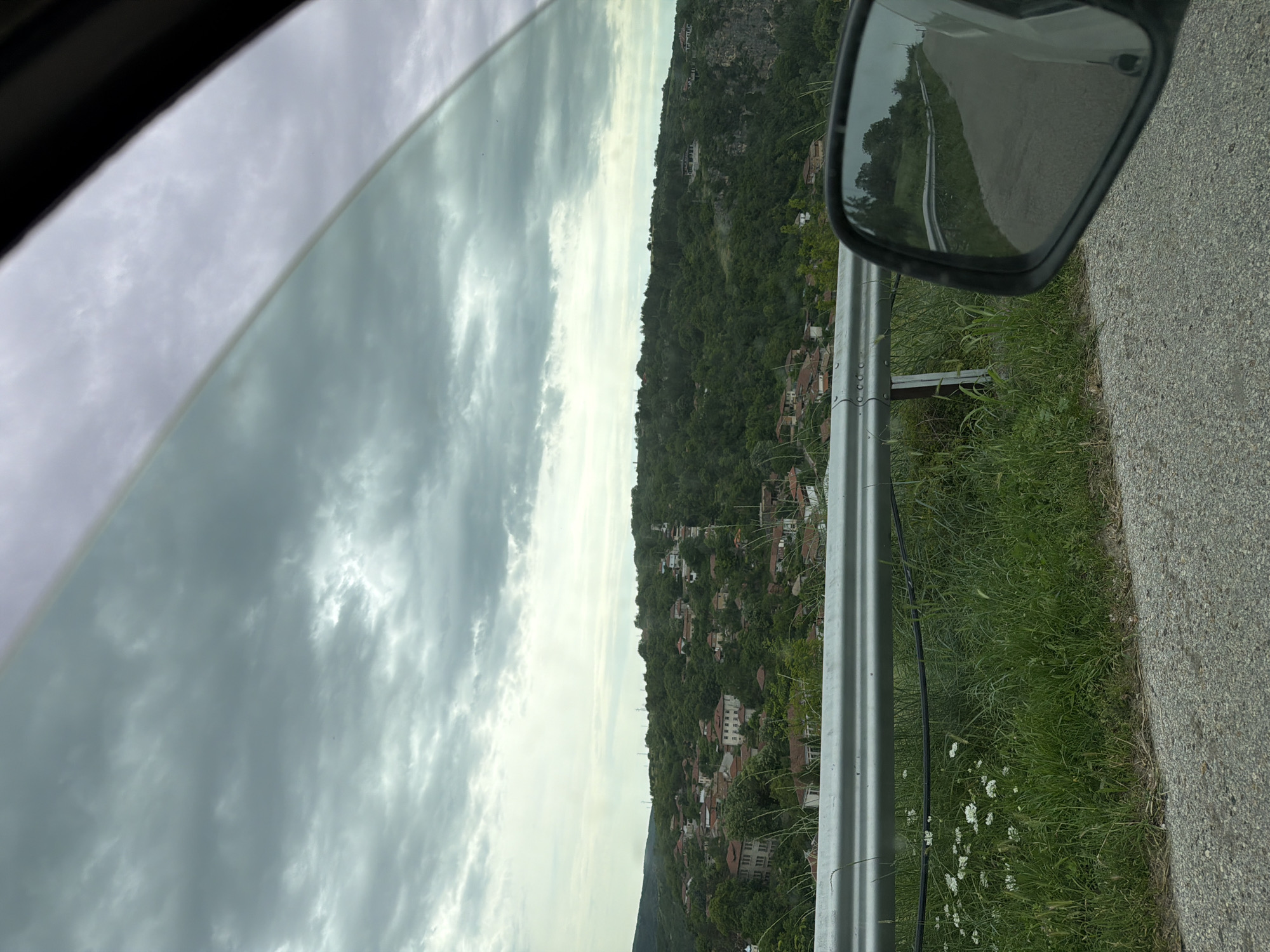 The image depicts a roadside scene in what appears to be the city of Cherven, Bulgaria, observed from inside a vehicle. The view through the car window reveals a cloudy sky occupying the upper left portion of the frame. The landscape features a hillside densely populated with trees and buildings, possibly residences, stretching along the center. A metal guardrail runs parallel to the road on the right, with grass and wildflowers growing along its base. The side mirror of the car is visible in the upper right, reflecting the road behind. The road surface is asphalt.