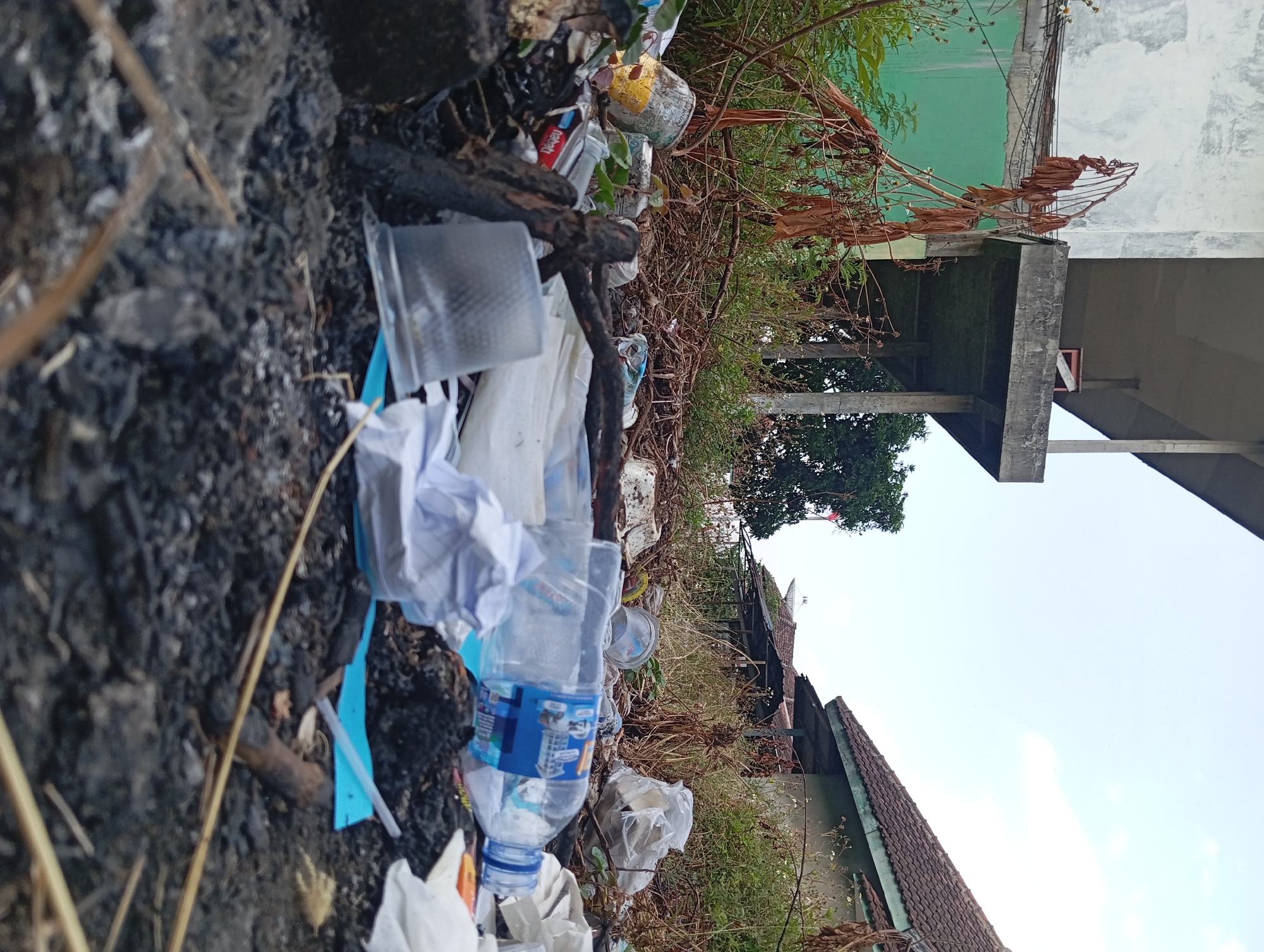 The image depicts a pile of garbage in front of an abandoned building. The garbage includes plastic bottles, paper, and other debris. The building appears to be in a state of disrepair, with peeling paint and overgrown vegetation. The sky is clear and the sun is shining. The scene is located in Pandaan, Indonesia.