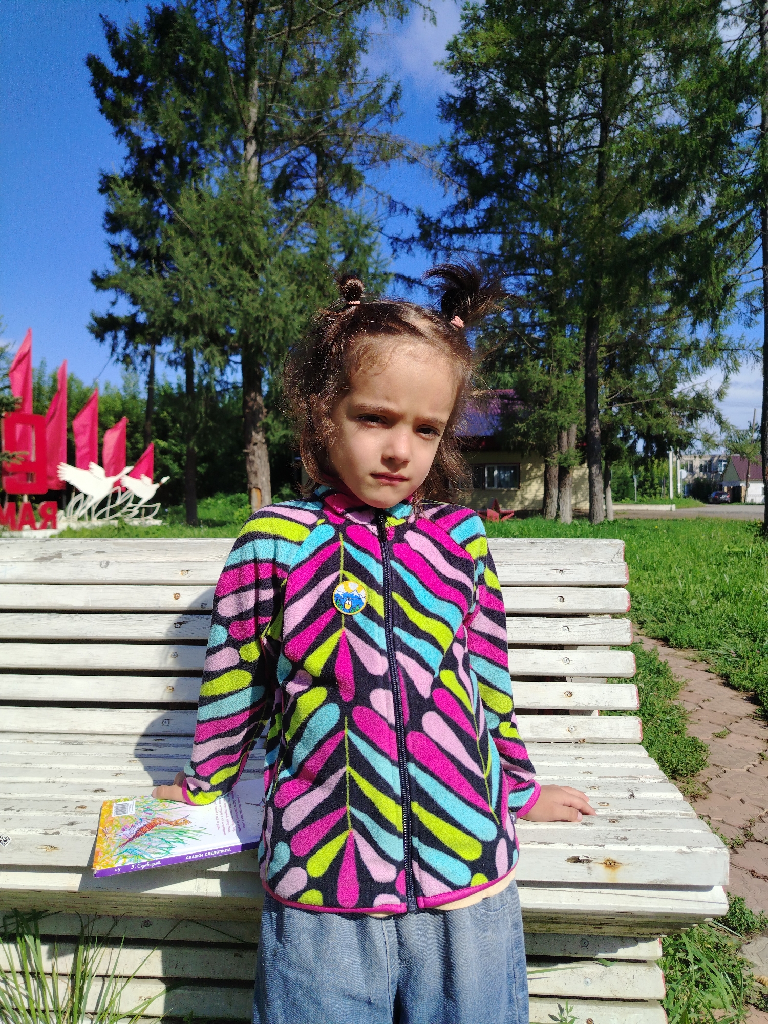 A young girl with dark hair styled in two buns is sitting on a white wooden bench outdoors. She is wearing a colorful fleece jacket with a zigzag pattern in pink, blue, green, and black, and blue denim shorts. A book titled "СКАЗКИ СЛЕДОПЫТА" (Tales of a Scout) is on the bench next to her. The background features tall evergreen trees, a clear blue sky, and some red flags and a white sculpture with two birds. In the distance, there are buildings and a road. This scene appears to be in the city of Kuyeda, Russia, possibly near a park or public space.