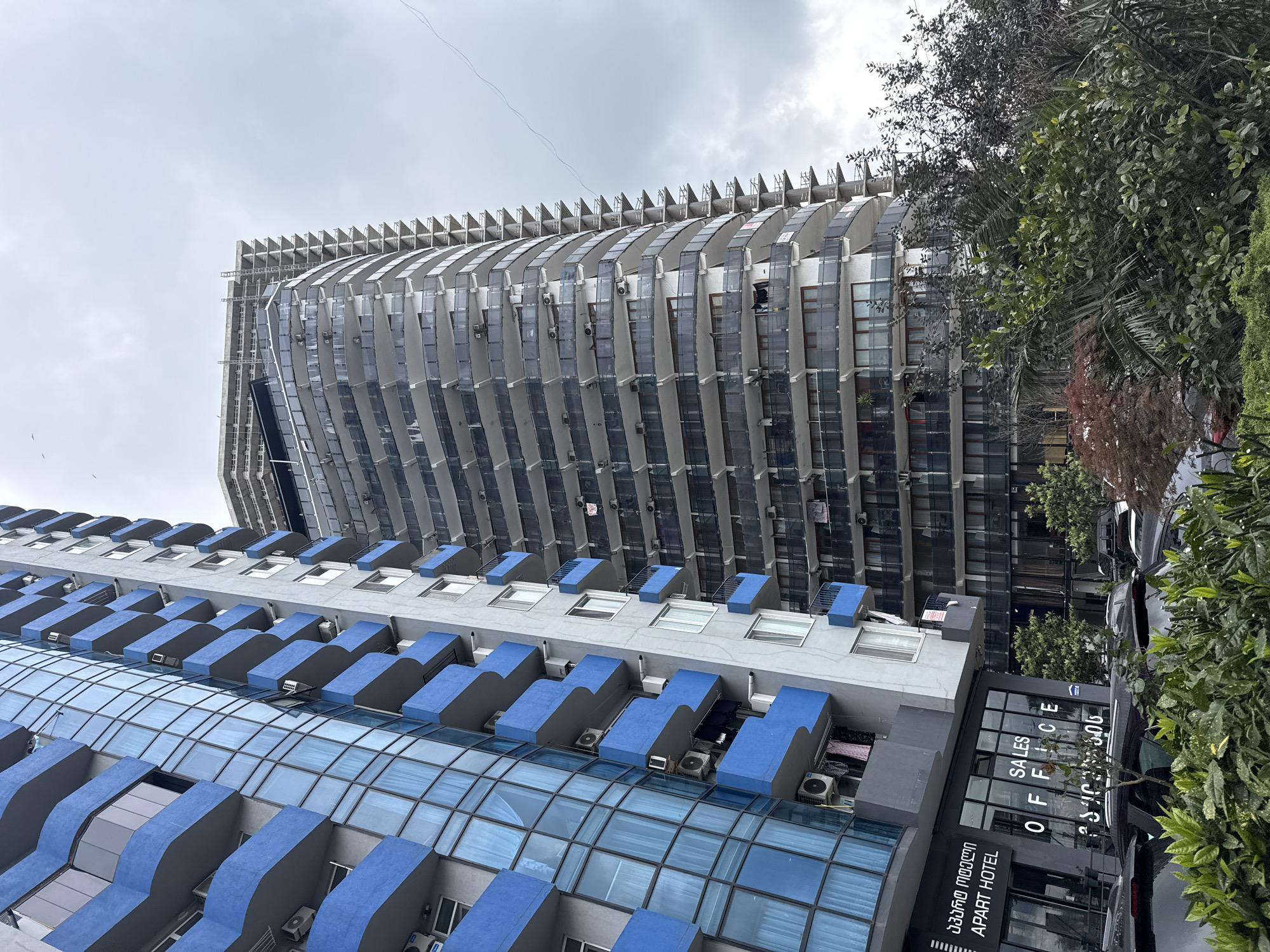 The image shows two modern buildings in Batumi, Georgia, under a cloudy sky.
The taller building has a curved facade with a series of protruding balconies. Many of these balconies have air conditioning units installed on them.
In the foreground, a lower building features blue balcony overhangs. A sign reads "SALES OFFICE APART HOTEL" in English and Georgian. A car is parked next to the building. Trees and vegetation are present on the right side of the image.