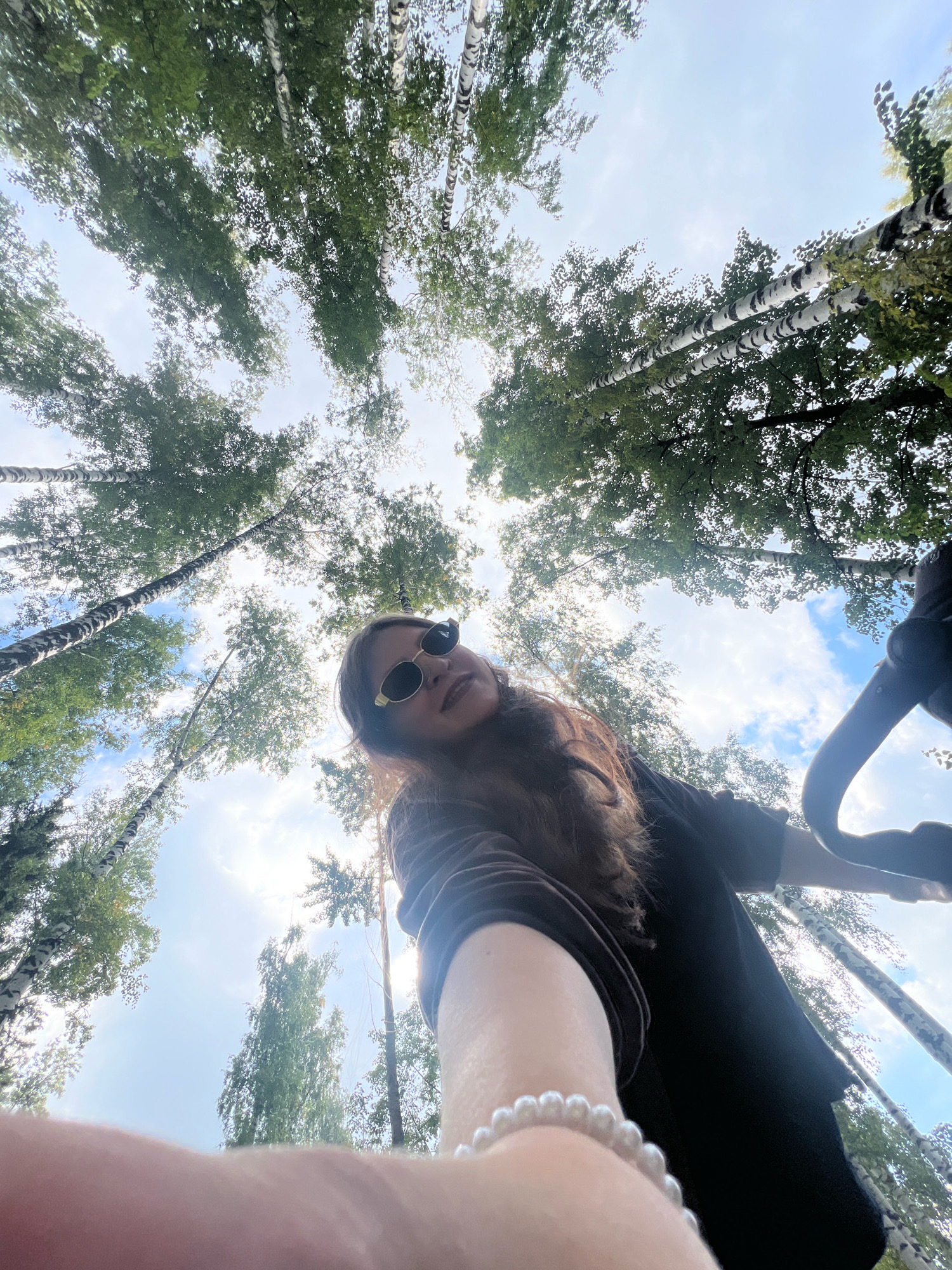 The image shows a low-angle, wide shot of a woman with long brown hair and sunglasses posing for a selfie in a wooded area. She is wearing a dark-colored top and a pearl bracelet on her left wrist. The background is dominated by the sky and tall, thin birch trees with white bark and green leaves. The sun is shining, creating bright highlights and dappled light filtering through the leaves. The scene appears to be outdoors in a natural setting, likely a park or forest, given the presence of birch trees. While the image's metadata does not specify a precise location within Noviy, Russia, the presence of birch trees strongly suggests a northern European or Siberian environment, consistent with that region.