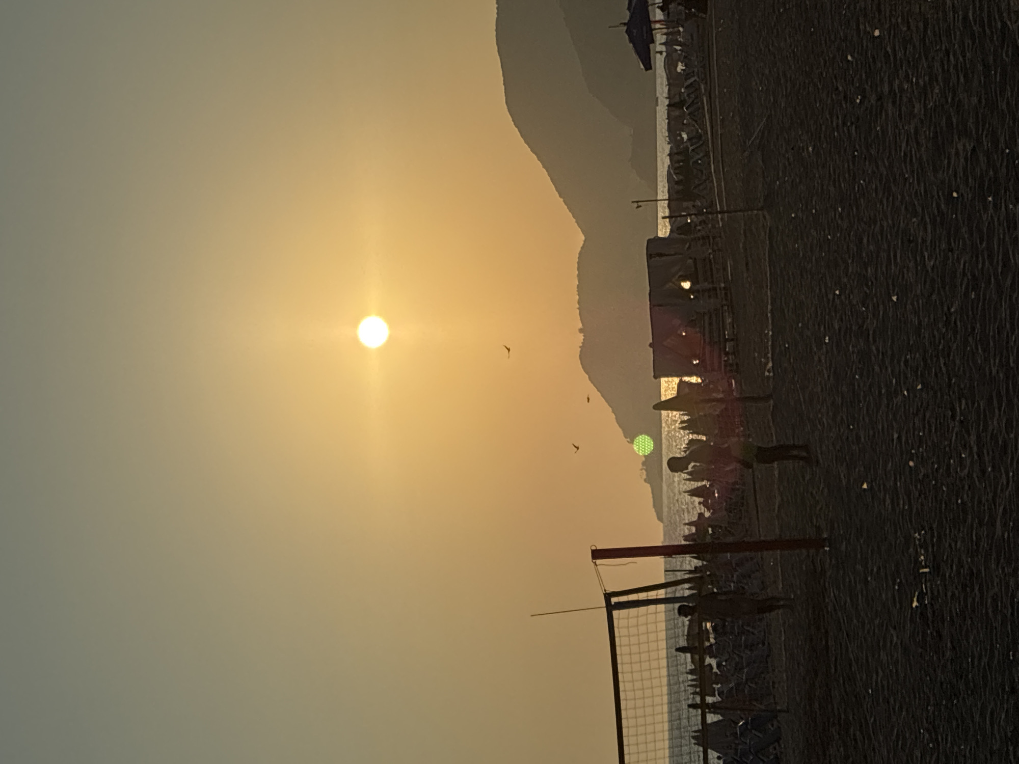 The image depicts a sunset scene on a beach in Alanya, Turkey. The sun, a bright orb with a halo, is low in the hazy, orange-yellow sky, casting a warm glow over the landscape. Silhouetted against the fading light are several people playing beach volleyball. A net is set up, and figures are visible on either side, suggesting a game in progress. In the background, the dark shapes of mountains rise above the sea, with a hint of buildings or structures along the coastline. The beach itself is dark, appearing to be made of sand or small pebbles. The overall mood is one of tranquil leisure as the day winds down. No visible text is present in the image.