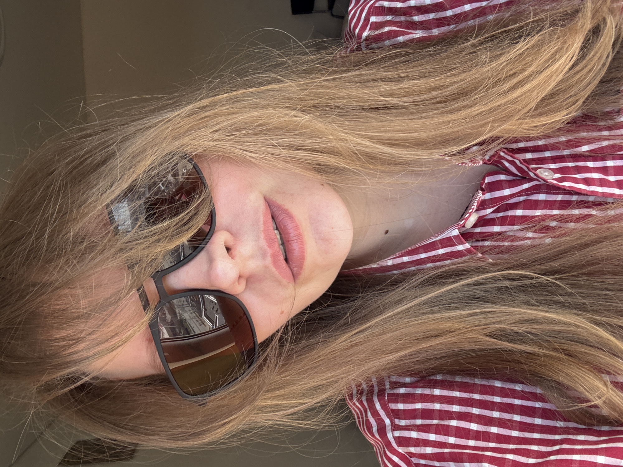 A woman with long blonde hair, wearing dark sunglasses, is shown from the chest up in an indoor setting. She is wearing a red and white striped shirt. The reflection in her sunglasses appears to show an urban setting, specifically architecture that could potentially place the image in Izhevsk, Russia. The angle of the image suggests it was taken from a tilted perspective. There are no additional people or specific objects clearly visible.