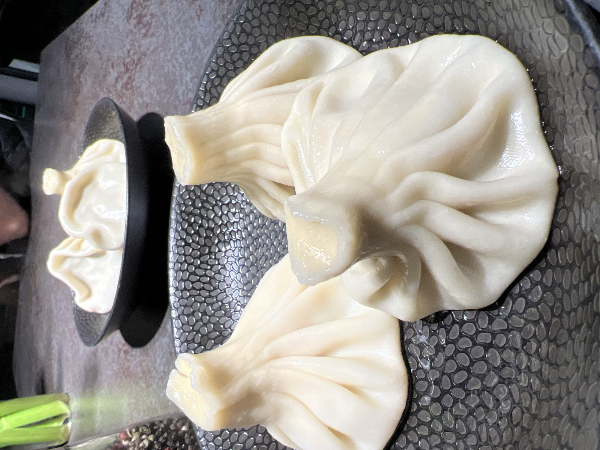 The image presents a close-up view of khinkali, a Georgian dumpling, arranged on two decorative plates. The first plate, a small black dish, contains one khinkali. The larger plate, featuring a textured gray and black pattern, holds two khinkali. A partial view of a glass container filled with green celery stalks and peppercorns is visible at the bottom left of the frame. Given the presence of khinkali and celery, it is plausible that the location is a restaurant, although the image does not confirm this. The location of this image in Budva, Montenegro, is not discernible from the provided visual details.