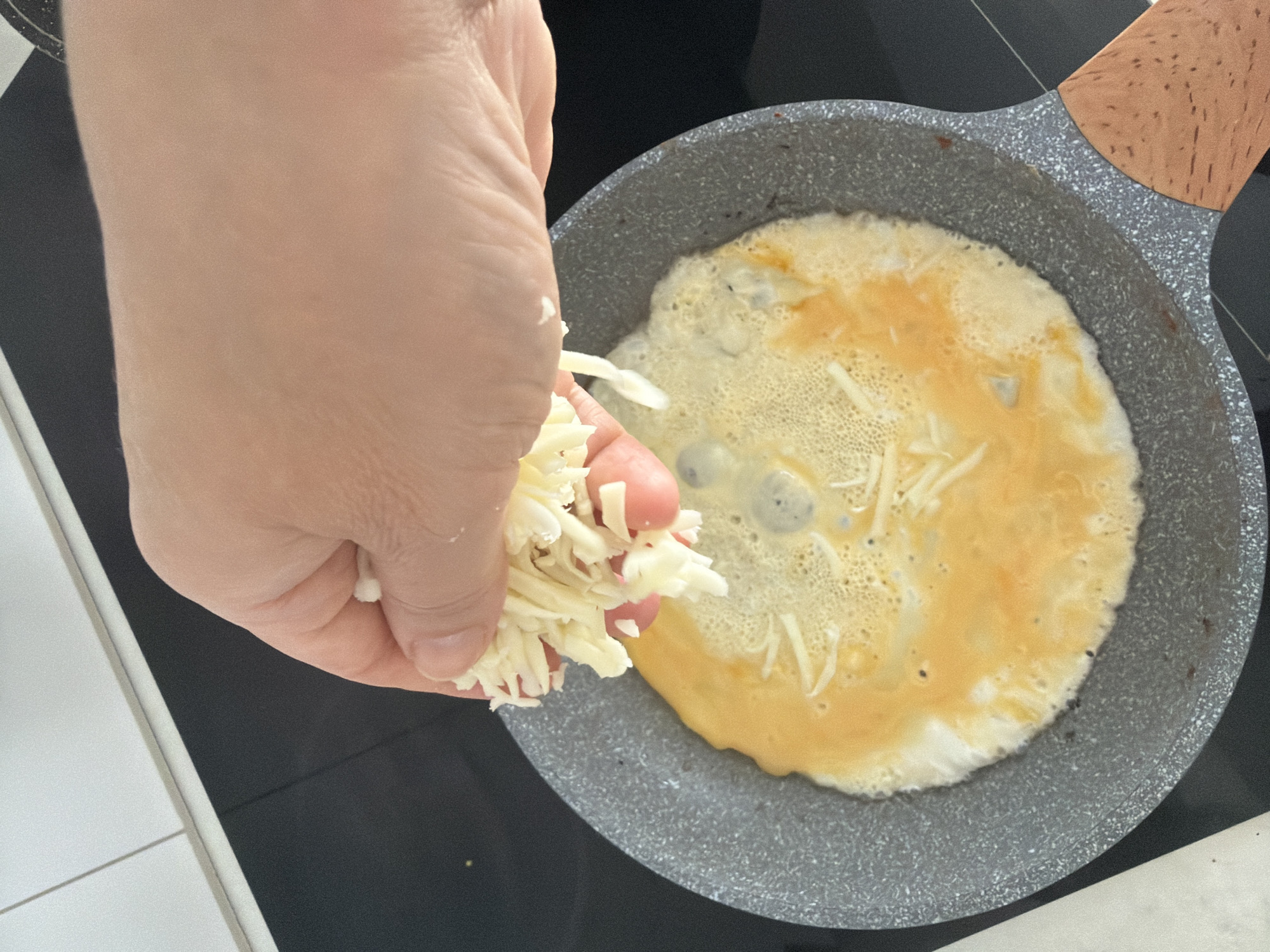 A close-up shot shows a hand sprinkling shredded cheese onto an omelet cooking in a non-stick frying pan. The omelet is partially cooked and golden brown, with some bubbles on its surface. The frying pan has a speckled gray interior and a light brown wooden handle. The pan is placed on a black induction stovetop. In the background, a white countertop with subtle tile lines is visible. This scene is taking place in Malinovka, Russia.