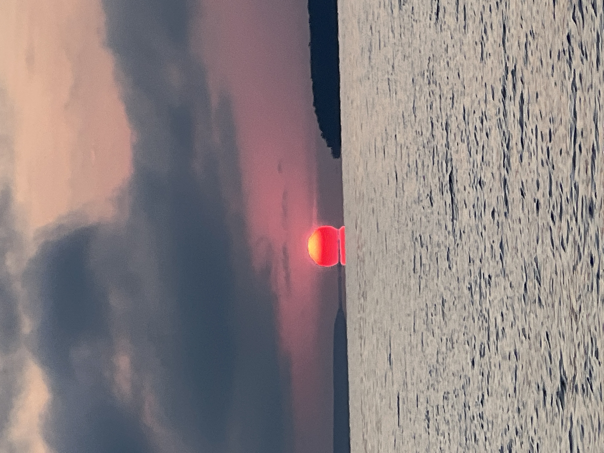 A circular, bright orange-red sun is partially submerged in the horizon of a body of water. The sun is reflecting on the surface of the water, creating a bright patch. Dark, heavy clouds occupy the upper half of the image, with a hint of pinkish-orange light peeking through the edges. A distant, dark landmass is visible on the right side of the horizon. The foreground consists of rippling water, reflecting the colors of the sky. The scene depicts a sunset over a lake or sea, possibly near Novopichugovo, Russia. There are no people or specific objects identifiable in the image.