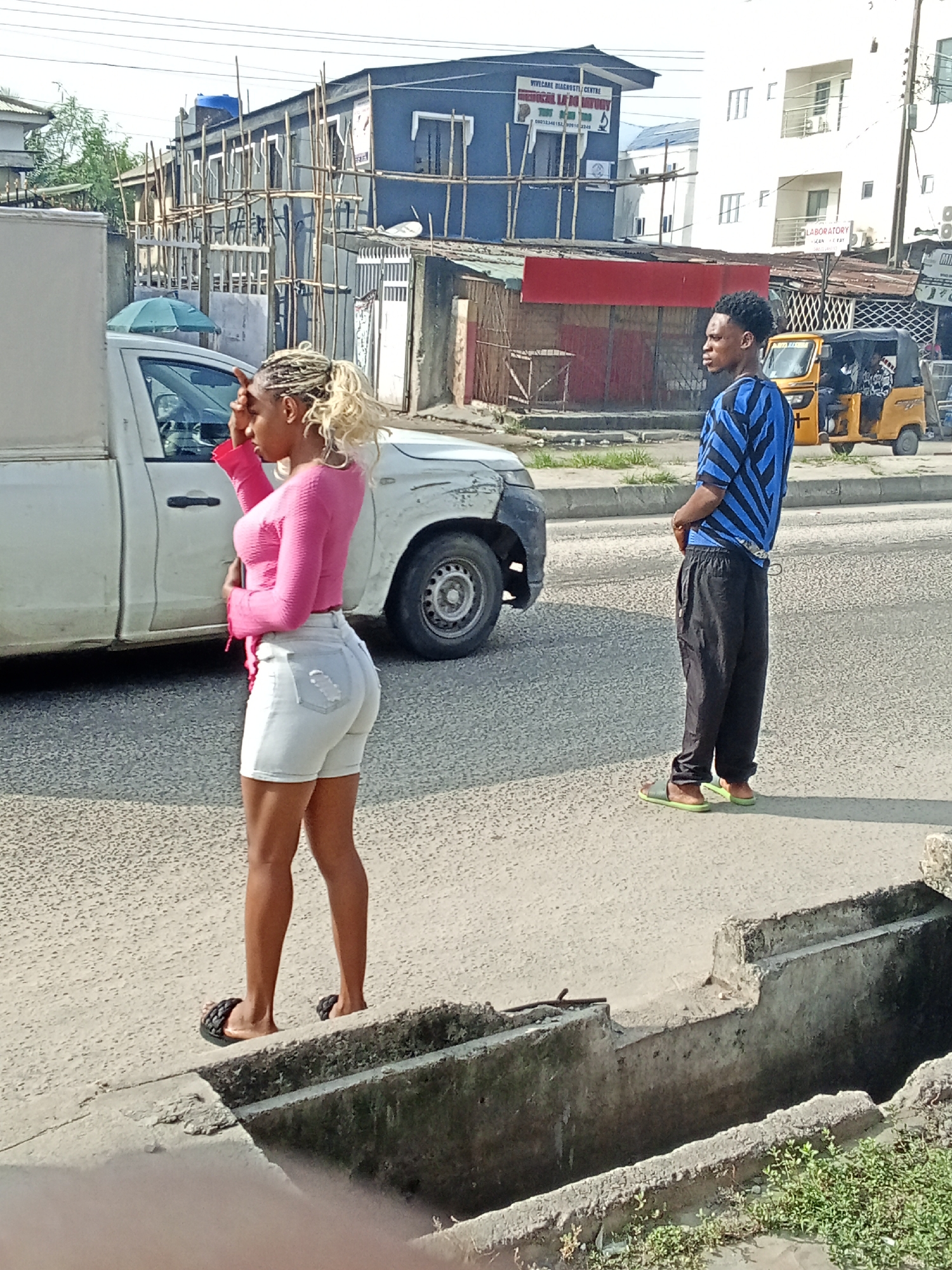 A street scene in Ado, Nigeria. A woman in a pink long-sleeved shirt and light-colored denim shorts stands on the edge of the sidewalk next to a white truck, looking to her left while touching her forehead. A man in a blue and black patterned shirt and black pants stands on the road, looking left. In the background, there is a yellow auto-rickshaw, buildings, and street signs. A concrete drainage ditch is in the foreground.
