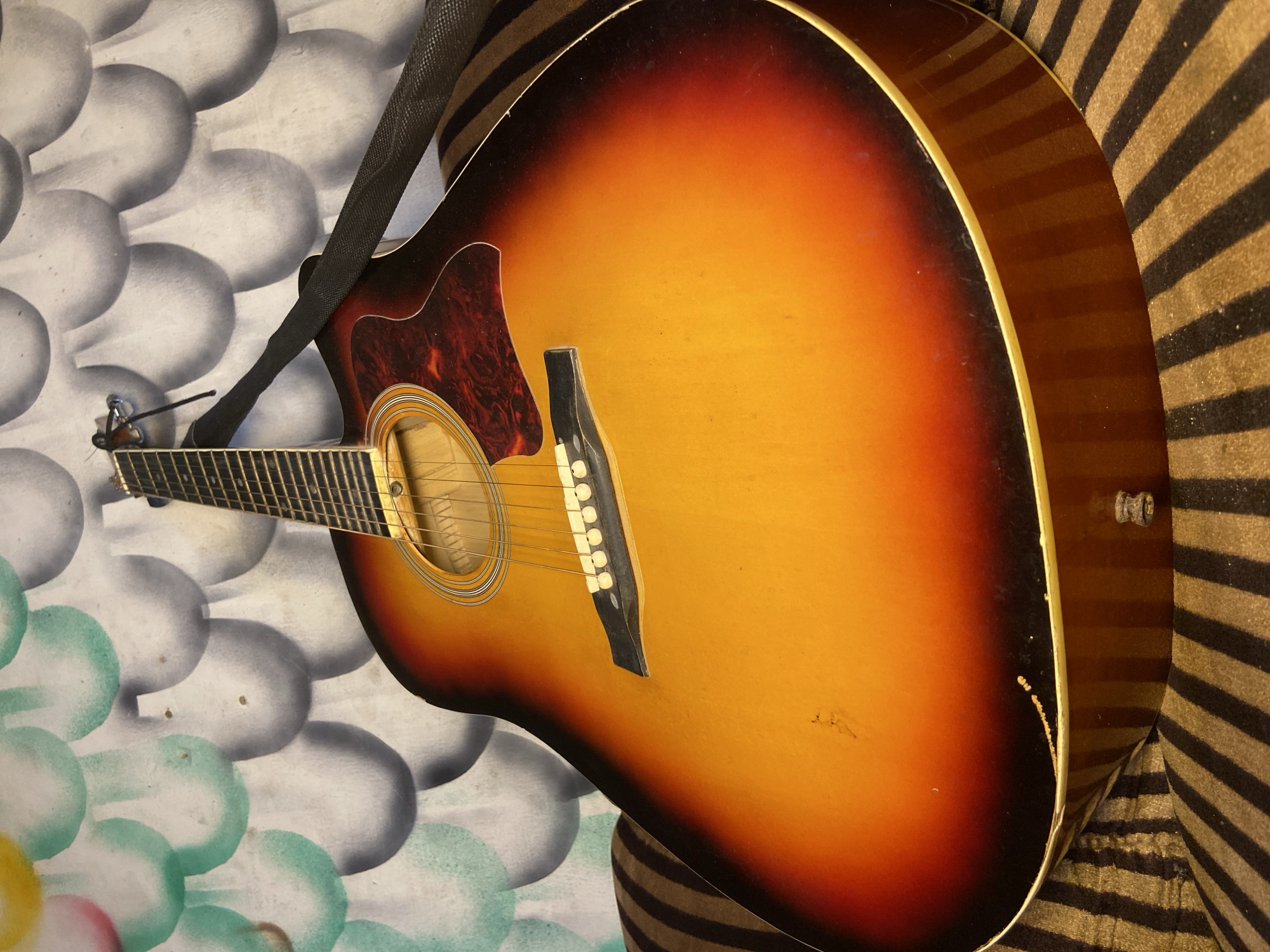 An acoustic guitar with a classic sunburst finish is the central subject, positioned in an indoor setting. Its body features a gradient from bright yellow-orange in the center to a dark reddish-brown and black around the edges. A dark, mottled reddish-brown pickguard is visible below the soundhole. Inside the soundhole, a label with the brand "YAMAHA" is faintly discernible. A black fabric strap is attached to the guitar, resting across its upper bout.
The background is divided into two parts. On the left, there's a wall decorated with a repeating pattern of large, overlapping ovoid shapes in shades of grey, white, and light green, with hints of other colors at the bottom. On the right, behind the guitar's lower body, a striped fabric is visible, consisting of alternating horizontal bands of dark brown/black and lighter brown/beige, possibly a cushion or chair.
The guitar appears to be used, exhibiting minor scuffs on its body and slight wear along its light-colored binding. The image captures the instrument in a stationary state, suggesting it is either stored or on display. There are no people visible, and no specific activity is taking place. The scene is illuminated by ambient indoor lighting. This media file originates from Mararaba, Nigeria.
