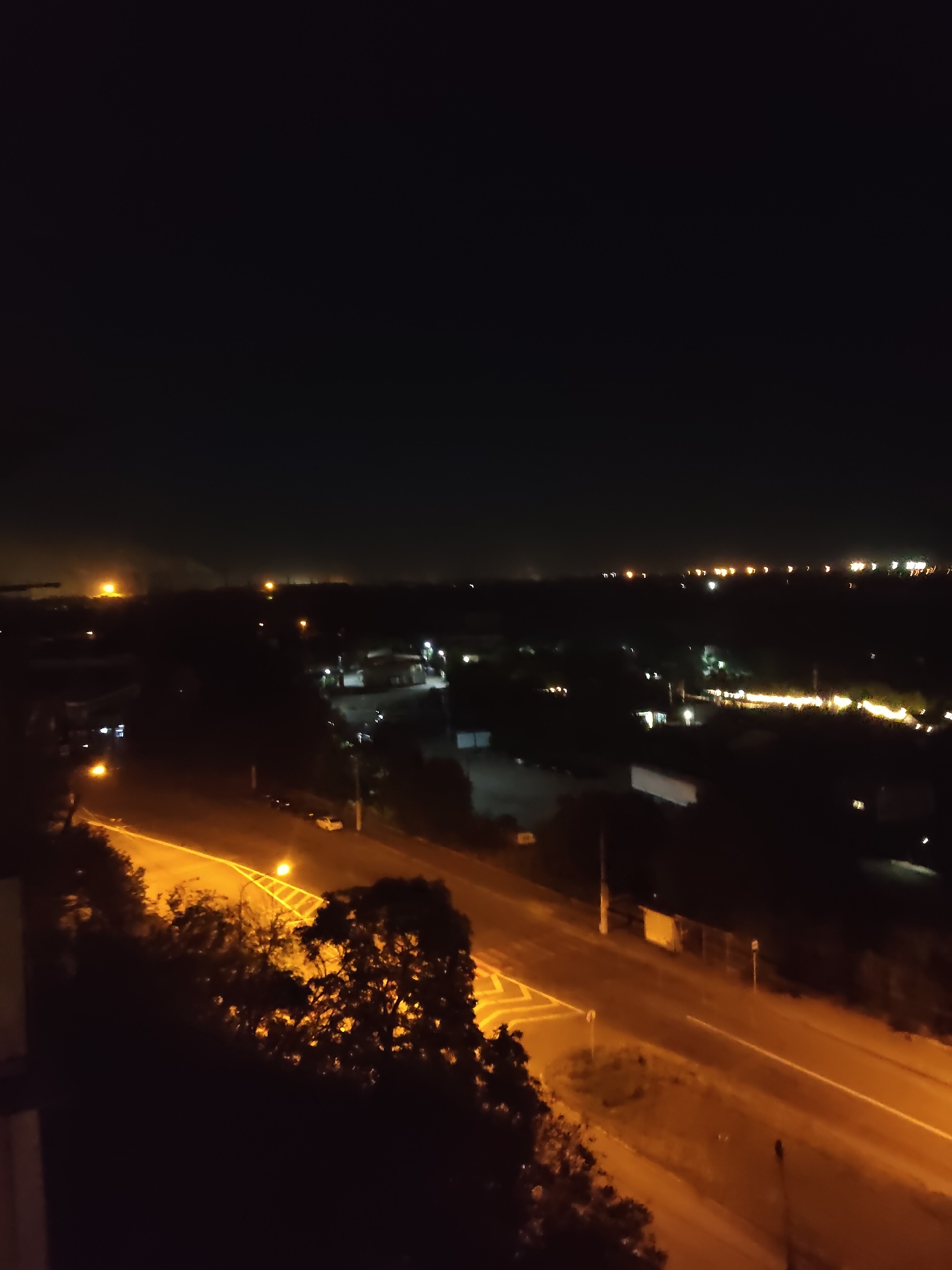 An aerial nighttime view of an urban or suburban landscape in Zaporizhzhia, Ukraine, reveals a dark sky above a dimly lit environment. A road with yellow streetlights cuts through the scene, illuminating trees and some scattered vehicles. Buildings and structures are visible in the distance, appearing as clusters of lights against the darkness. The overall impression is one of a quiet night, with the artificial lights of the city providing the only significant illumination.