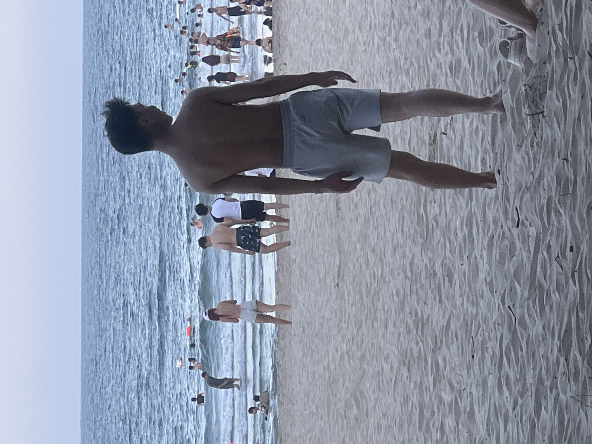 The image shows a crowded beach scene in Da Nang, Vietnam. In the foreground, a young man stands with his back to the camera, wearing light gray swim shorts. He is facing the ocean where many people are swimming and wading. The sand in the foreground is textured with ripples. In the background, the ocean is visible with gentle waves and a hazy sky. Numerous individuals are scattered throughout the water, some closer to the shore and others further out. The overall impression is of a busy recreational area on a warm day.
