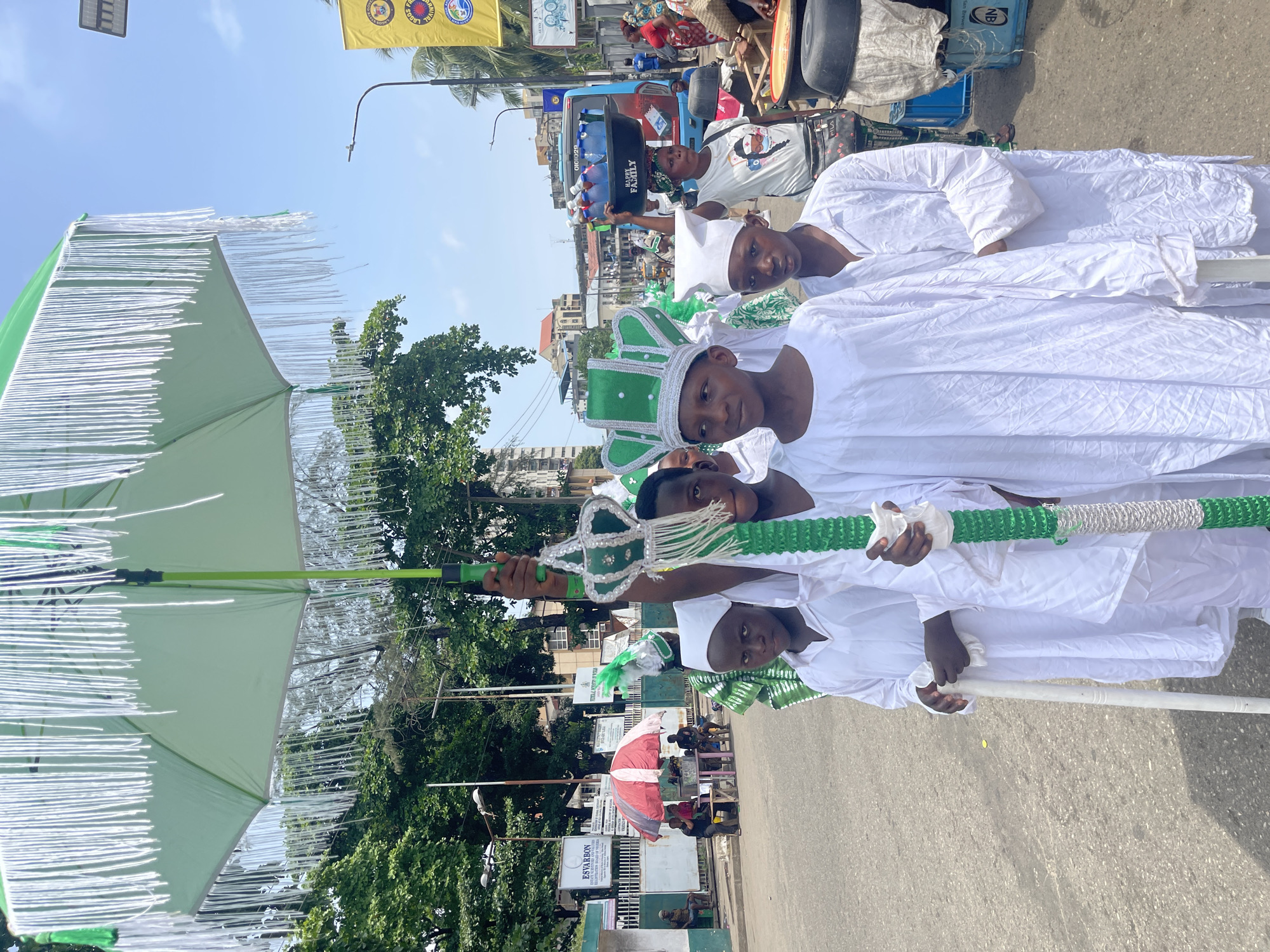 Four boys are dressed in white robes and white hats, with green and white trim, on a street. One of the boys is holding a large green and white umbrella and another is holding a green and white staff. A person sits in a car in the background and many trees are visible. A green sign reads "ESVARBON" and "REGISTRATION REQUIRED BEFORE ENTRY". The location is likely in Lagos, Nigeria, based on the green and white colors, resembling the Nigerian flag.