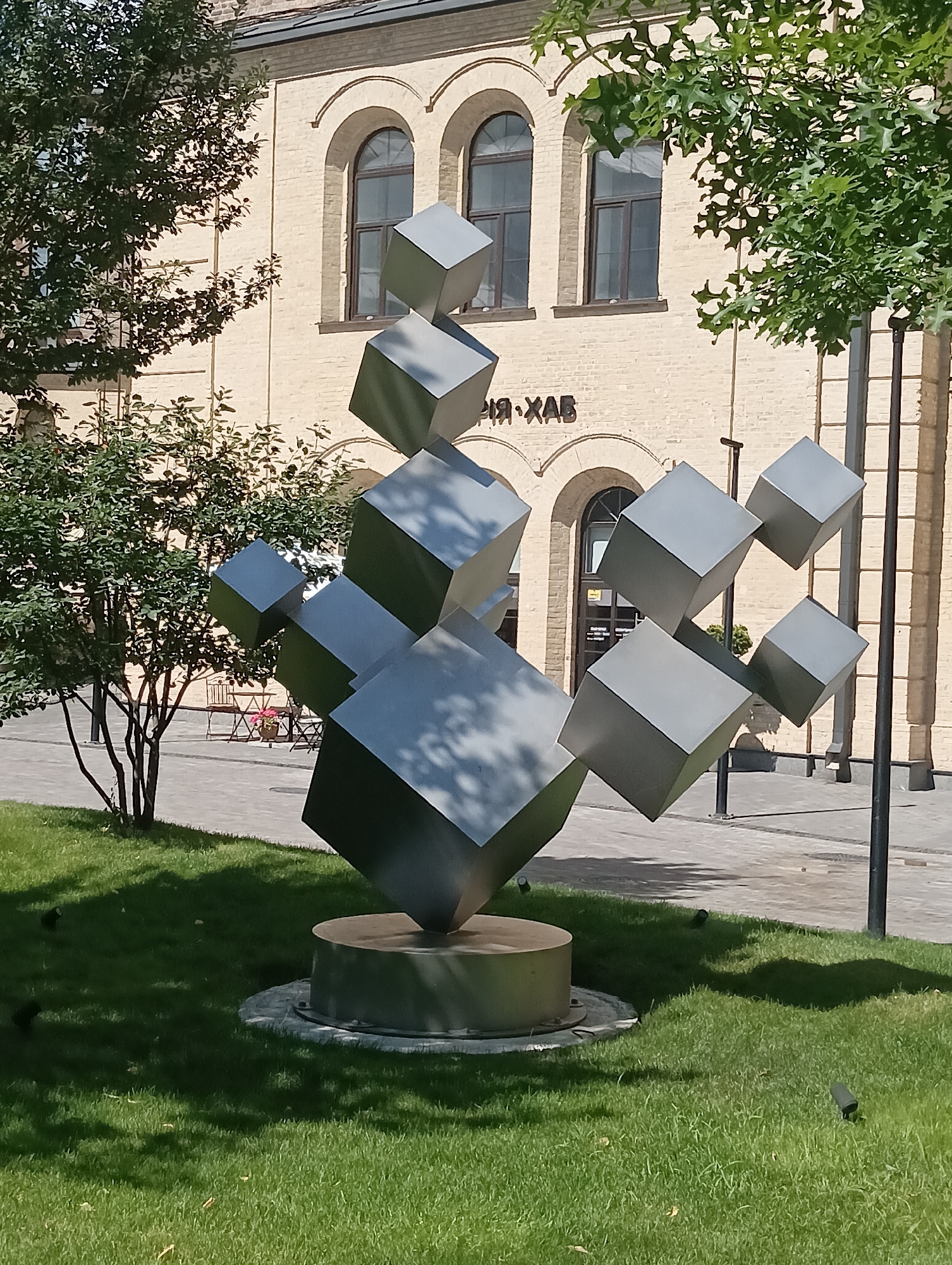 A modern sculpture of interlocking cubes stands on a circular stone base on a green lawn in front of a brick building. The cubes are made of a smooth, gray material and are arranged in a dynamic, abstract formation. Some of the cubes are angled and appear to be floating in mid-air. The building behind the sculpture has arched windows and a light-colored brick facade. There are trees and bushes to the left of the sculpture, and a paved walkway is visible in the background. The scene is illuminated by bright sunlight, casting shadows on the lawn and sculpture. The overall impression is one of artistic expression and urban landscaping.