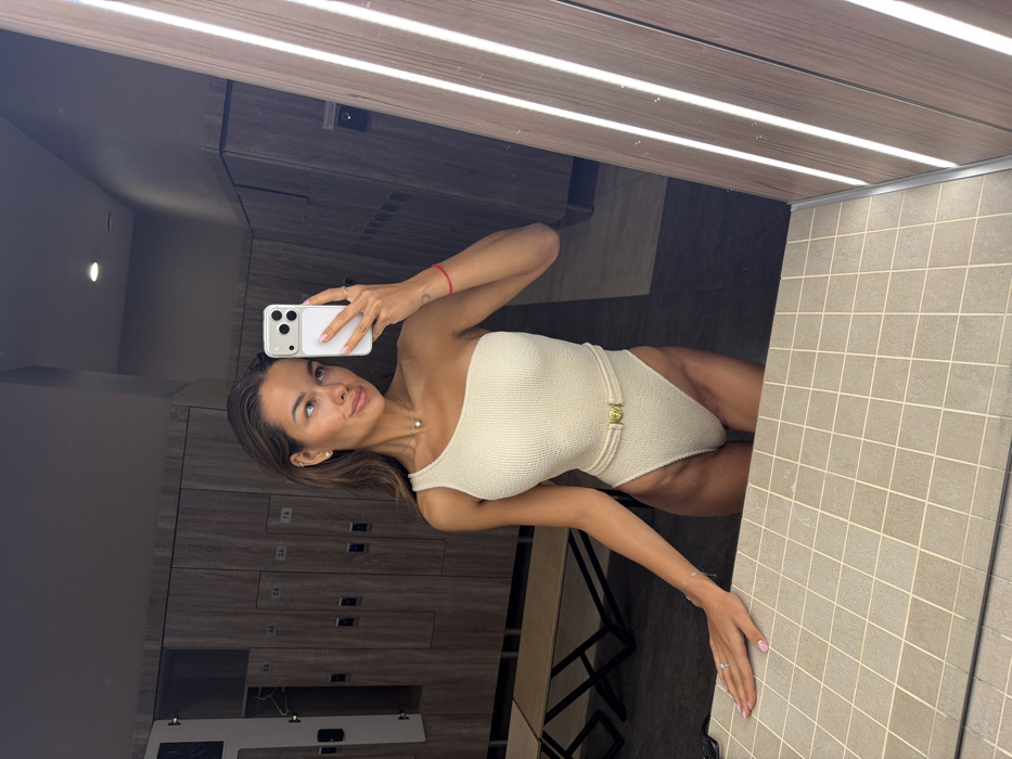 The scene depicts a modern changing room or locker facility, characteristic of a gym, spa, or aquatic center found in urban settings like Krasnoyarsk, Russia.
An adult female individual, appearing to be in her twenties or thirties, is featured taking a mirror selfie. Her dark brown hair is wet and styled back. She wears a light beige, textured, one-shoulder one-piece swimsuit, which includes a thin matching fabric belt with a gold-colored buckle around the waist. Her right hand holds a white-cased smartphone with a multi-lens camera system, angled to capture her reflection. Her left hand rests on the light grey tiled countertop in the foreground. She wears a small stud earring in her left ear, a delicate pendant necklace, and rings on her left thumb and ring finger, along with a red string bracelet on her right wrist. Her expression is a slight smile.
The background shows a row of light wood-tone lockers, several bearing visible numbers (e.g., 76, 78), with one on the far left slightly open to reveal a dark interior. Beneath the lockers, a light-colored bench with a dark metal frame is visible. The foreground is dominated by a countertop composed of square grey tiles with darker grout lines. Lighting includes a recessed ceiling light reflecting in the mirror and vertical LED strip lights integrated into the right side of the mirror frame. The overall environment is clean, well-lit, and possesses a contemporary aesthetic.