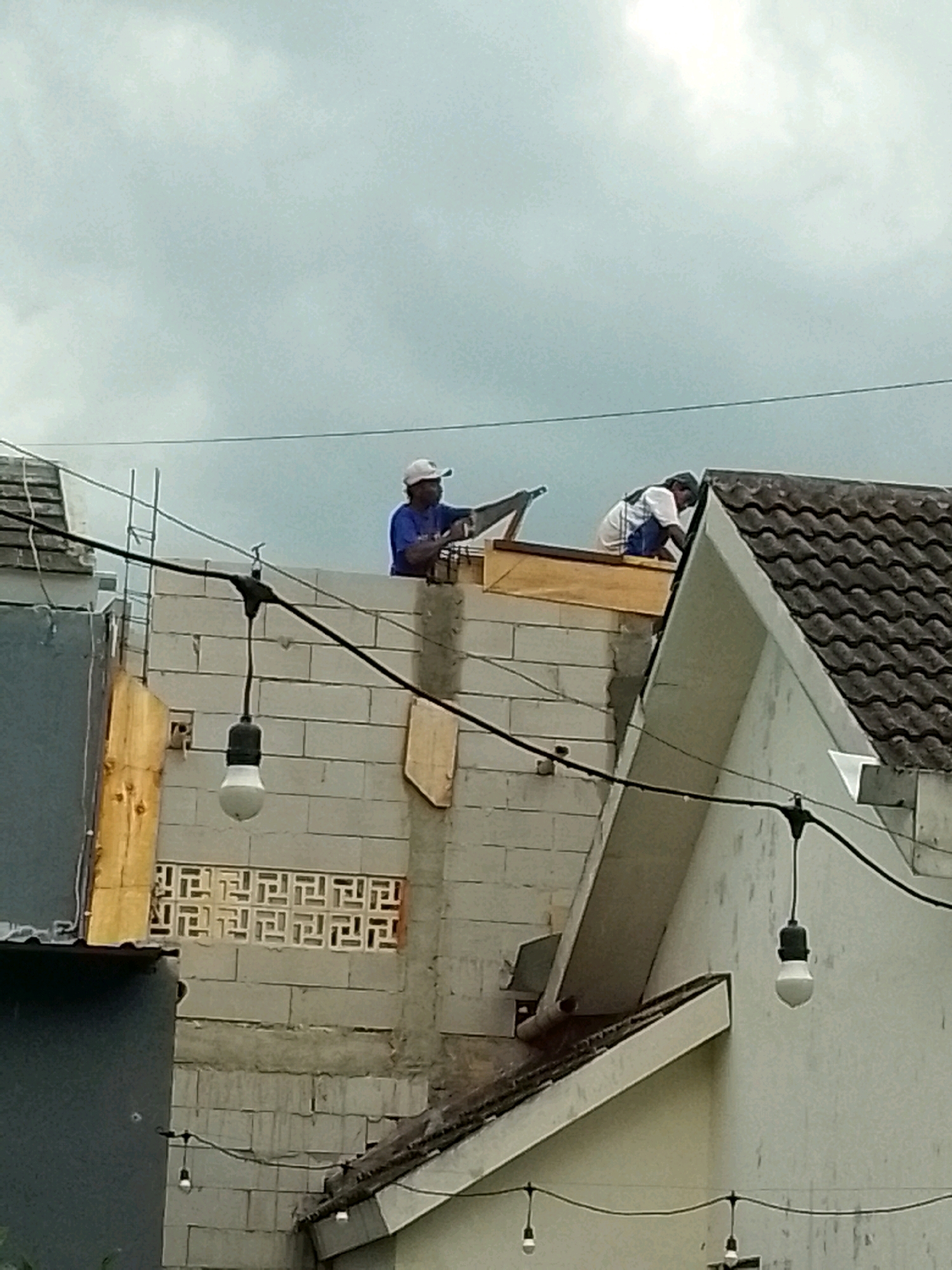 Two men are working on the roof of a building under construction in Jember, Indonesia. The man in the foreground, wearing a blue shirt and white cap, is holding a piece of wood. The second man, wearing a white shirt with blue accents, is crouched down, working on the roof. The building appears to be made of white bricks and has a tiled roof. There are wires and light bulbs hanging from the structure. The sky is overcast.