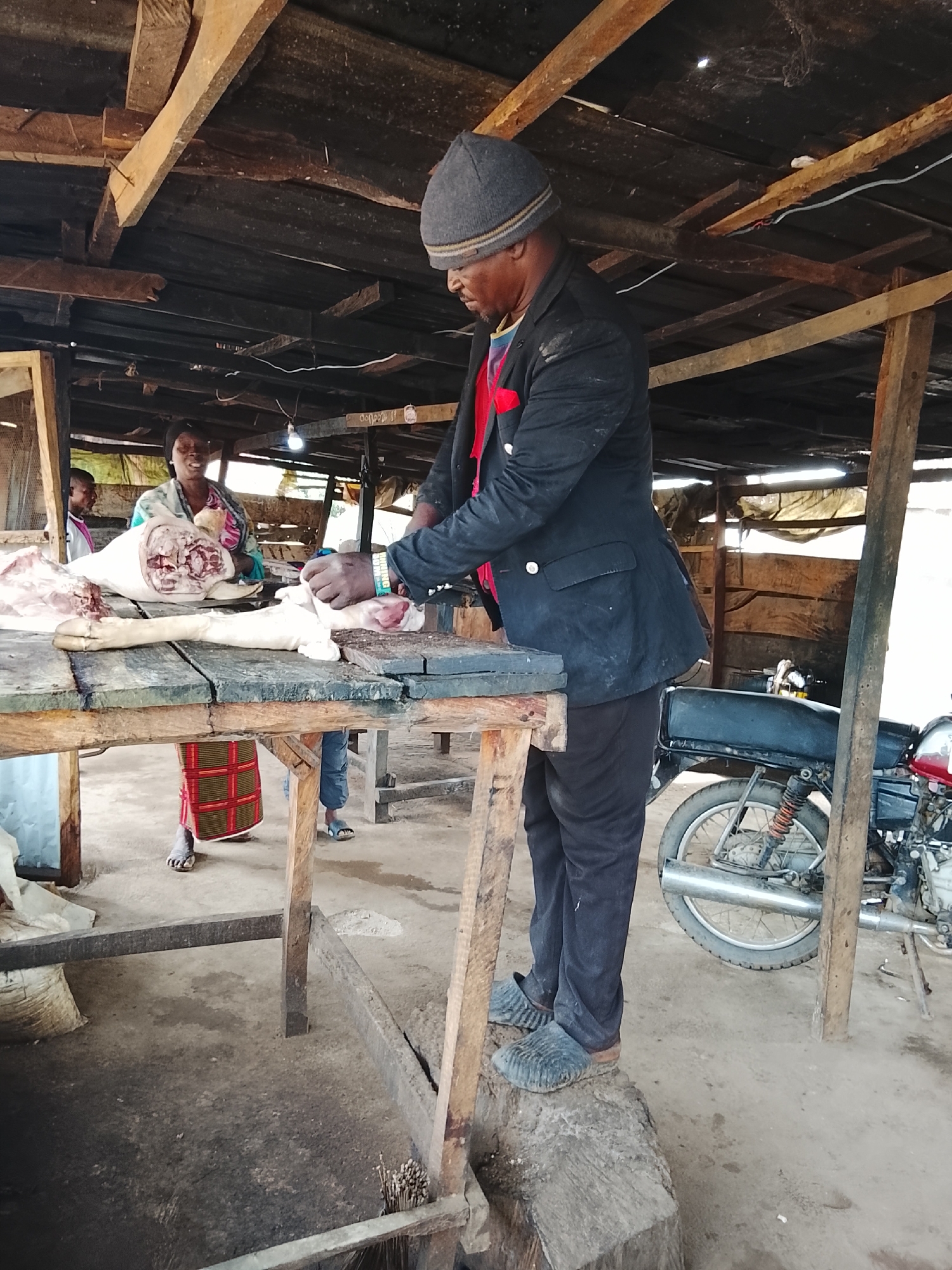 In Akwanga, Nigeria, a butcher, wearing a grey beanie, black blazer, red scarf, and black pants, stands on a wooden block to reach a table. He is in the process of butchering a pig carcass, with a knife in his right hand. A woman in a colorful top and a patterned skirt stands behind the table, holding part of the pig. Another person is visible behind her. The environment appears to be an outdoor market stall with a wooden roof supported by posts. A motorcycle is parked to the right of the scene. The ground is made of concrete.