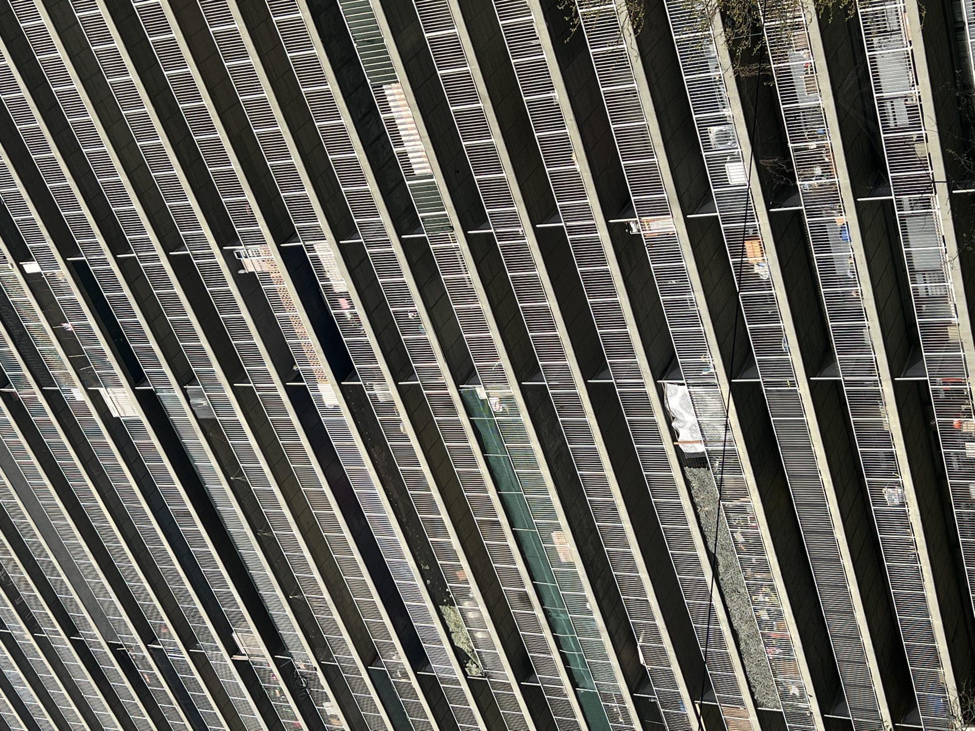 The image shows an aerial view of a high-rise building with multiple balconies, each featuring horizontal slats. These balconies appear to be external spaces for apartments or offices. The building has a repeating pattern of recessed areas between the balconies, giving it a striped appearance. The balconies contain various objects such as furniture, plants, and laundry, suggesting that they are in use. The photograph was taken in São Paulo, Brazil.