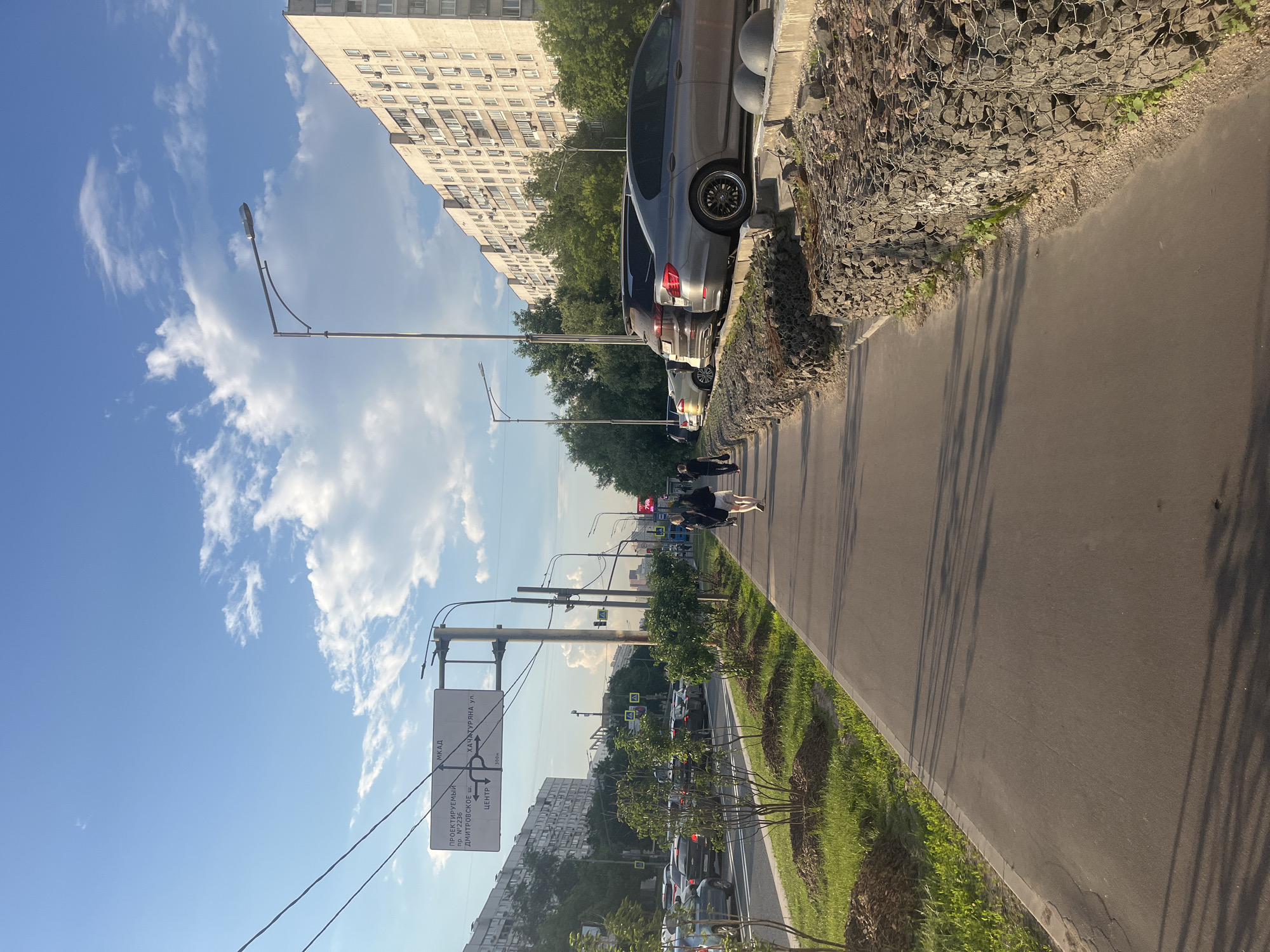The image shows a sidewalk adjacent to a multi-lane road in Moscow, Russia. The road is lined with parked cars and ongoing traffic. On the left side of the road, there are residential buildings and trees. On the right side of the sidewalk, there is a gabion wall filled with stones and some vegetation. Several people are walking on the sidewalk, moving away from the camera. A street light and traffic signs are visible. The sky is blue with scattered clouds, and the sun is casting shadows.