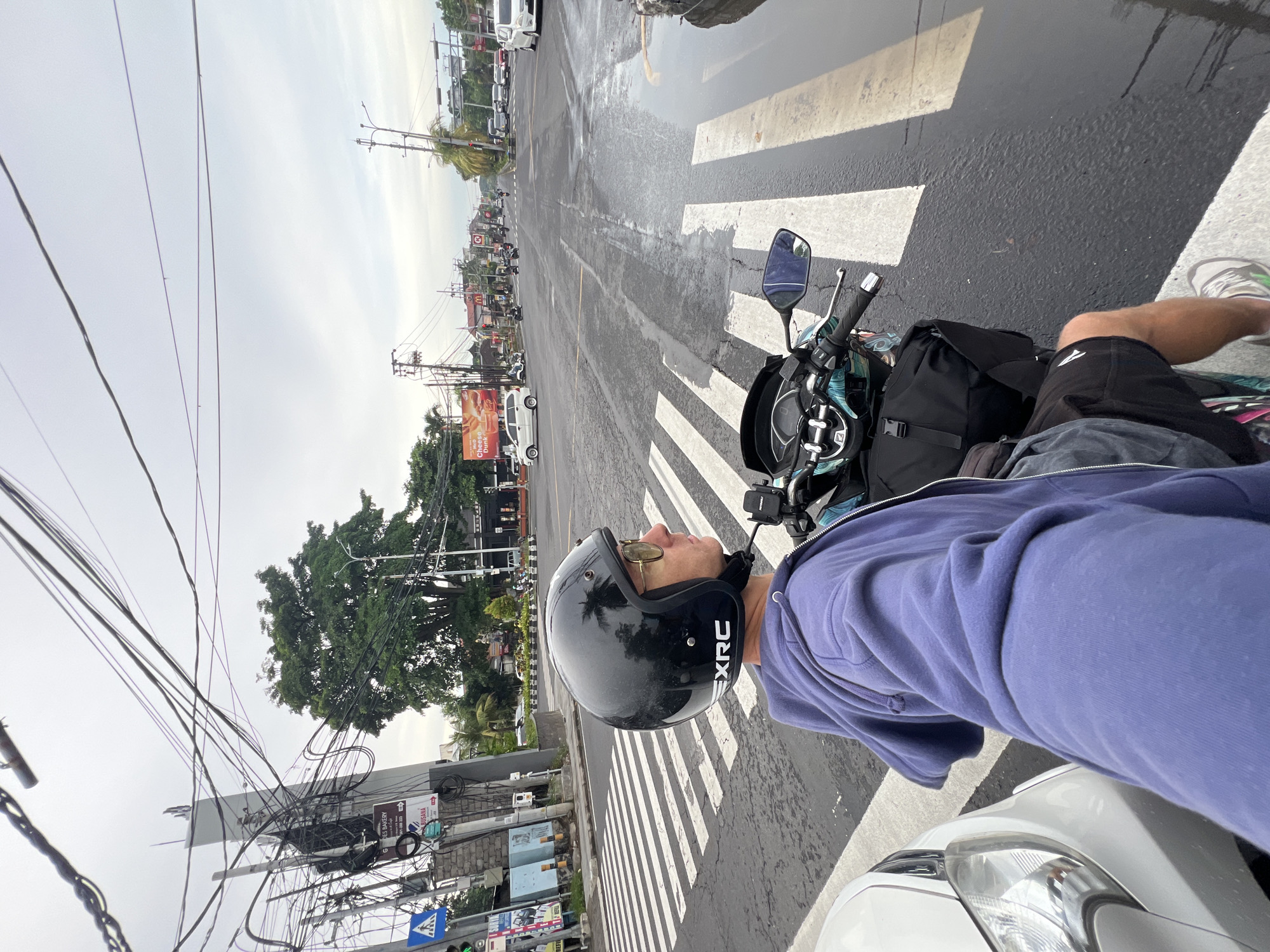 A man wearing a black EXRC helmet and a blue jacket is on a motorcycle at a crosswalk. The wet road surface indicates recent rain. He's wearing sunglasses. The location is an urban area, identified as Denpasar, Indonesia, based on the architecture and signage. Power lines are visible overhead, a common feature in the area. A restaurant is visible, including a McDonald's restaurant with a sign depicting a cheeseburger. Cars and other vehicles are on the road.
