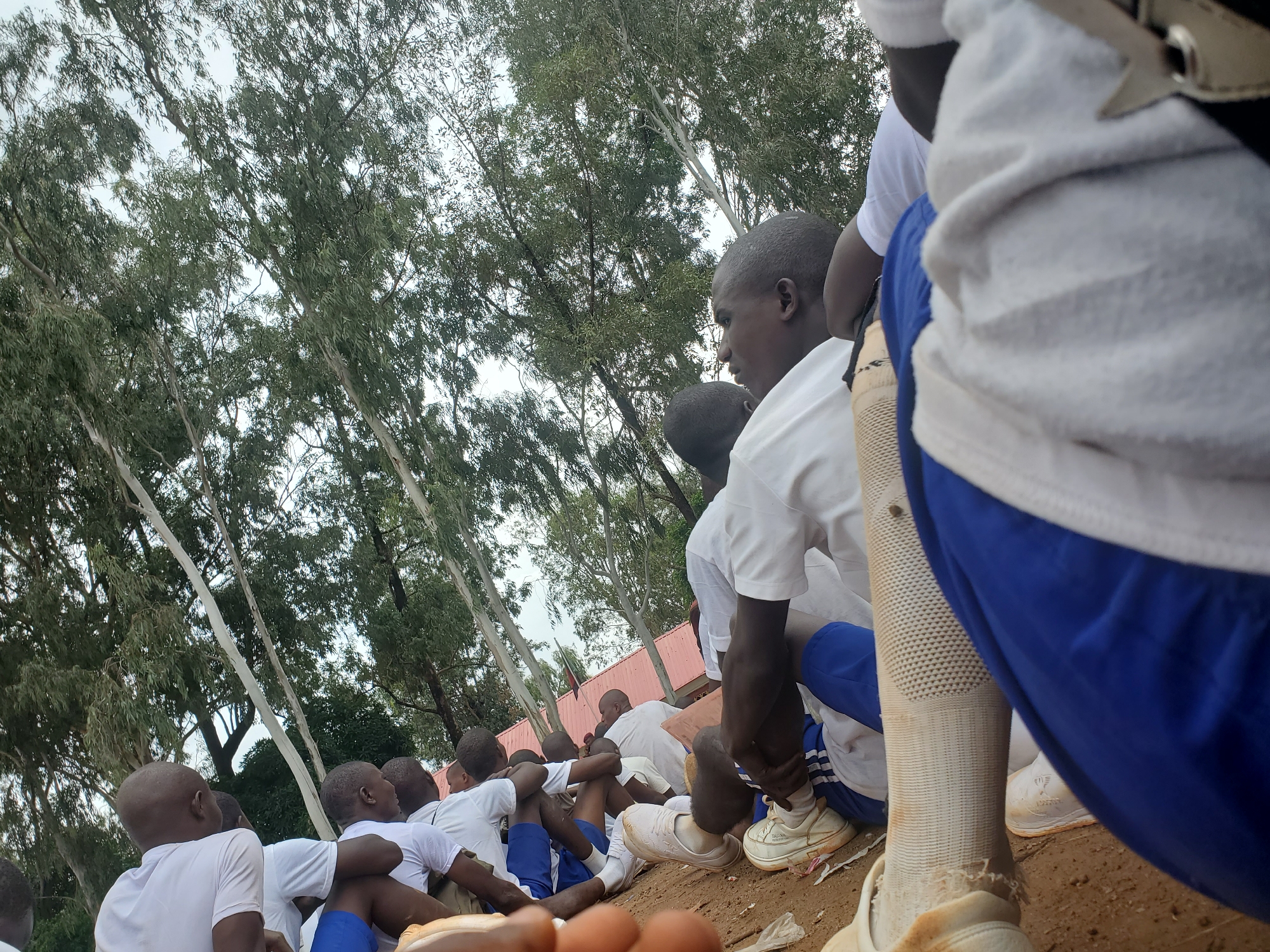 The image depicts a group of young men, seemingly in a training or exercise setting, outdoors. They are dressed in white t-shirts and blue shorts or athletic pants. Some are sitting on the ground, others are in a resting or stretching pose. The environment appears to be a dusty or sandy area, with trees and foliage in the background, suggesting a rural or semi-urban location. The overall scene conveys a sense of physical activity and camaraderie. The text references "Bahit, Nigeria" as the location, implying this is a specific outdoor area within that Nigerian city.