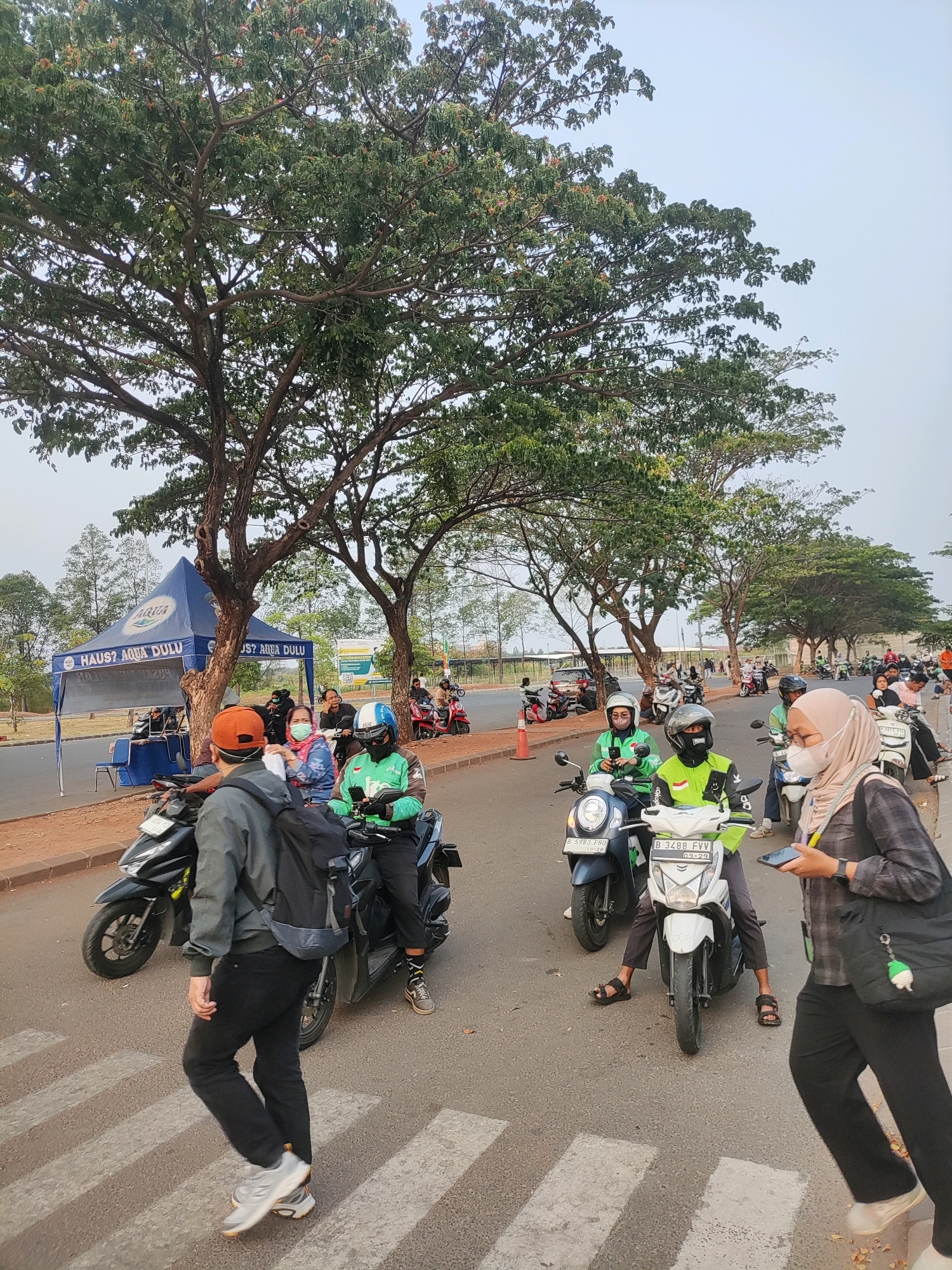 The image depicts a street scene in Telagamurni, Indonesia, featuring a procession of motorbikes and pedestrians. A large tent, likely for an event or vendor, stands on the left side of the road, displaying the "AQUA" logo and text that appears to read "HAUS? AQUA DULU". Several individuals are seated or standing around the tent. A group of people on motorbikes are moving along the road, some wearing helmets and branded green jackets, suggesting they might be delivery or ride-sharing service personnel. Pedestrians are also present, including one person in the foreground walking across a pedestrian crossing and another woman on the right side of the road holding a smartphone. The environment appears to be a public street with trees lining the background and a paved road. The sky is overcast.