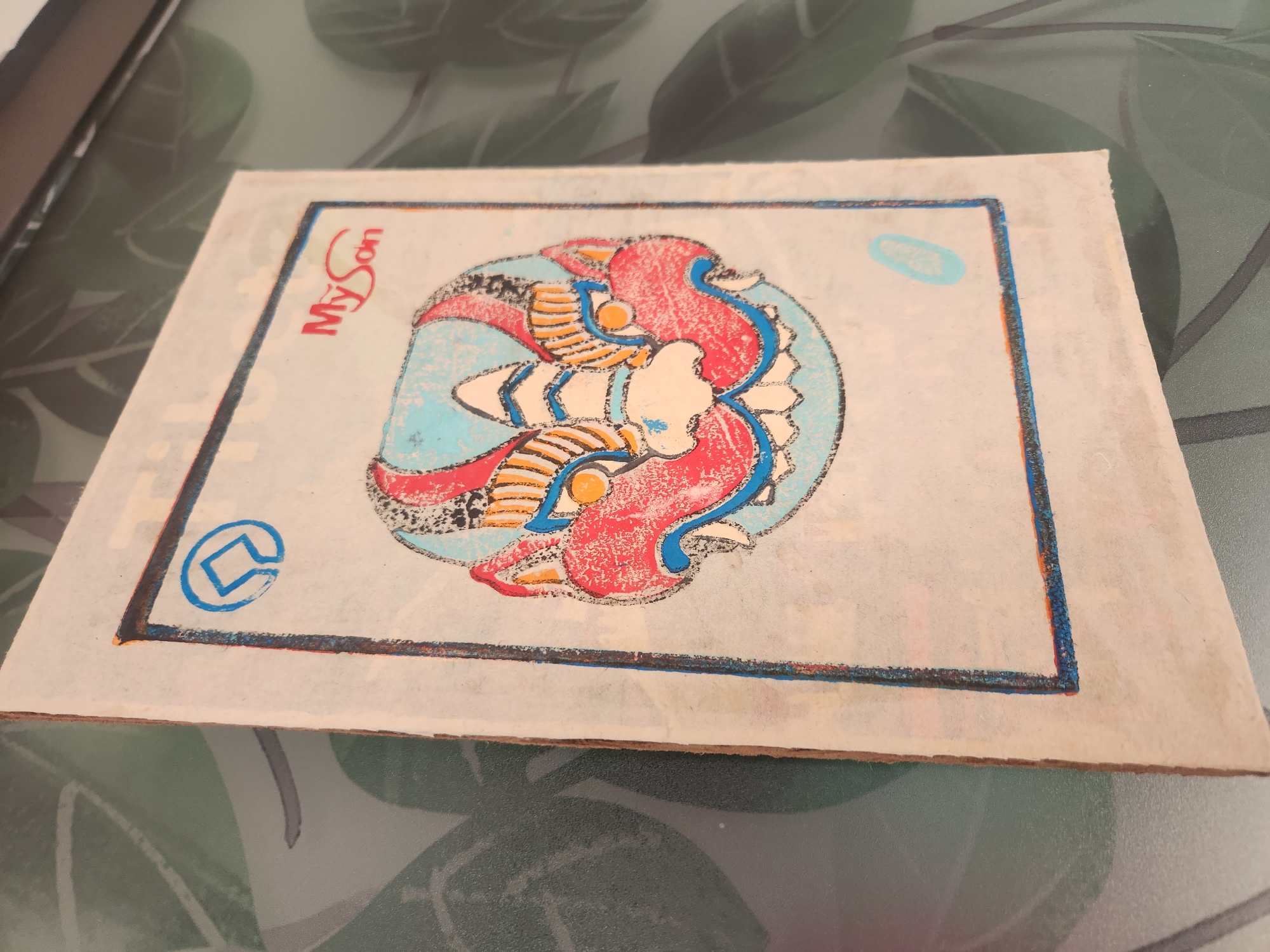 The image shows a close-up, eye-level, full shot of a piece of artwork featuring a stylized, colorful depiction of a mythological creature's face, possibly a dragon or a lion-dog, within a bordered frame on what appears to be handmade paper. The creature's face is predominantly red and blue with yellow and orange accents, characterized by sharp teeth, glowing orange eyes, and an aggressive expression. At the top left corner of the frame is a blue circular logo with a square inside. To the right of the logo, the text "MySon" is written in red cursive. The artwork is placed on a surface with a green, leafy pattern, possibly a table or a mat, and is angled to show its side edge, revealing a rough, layered texture of the paper. The background is out of focus, displaying blurred green and grey tones suggestive of a patterned surface. There is no discernible location in the city of Đà Nẵng, Vietnam, visible in the image.