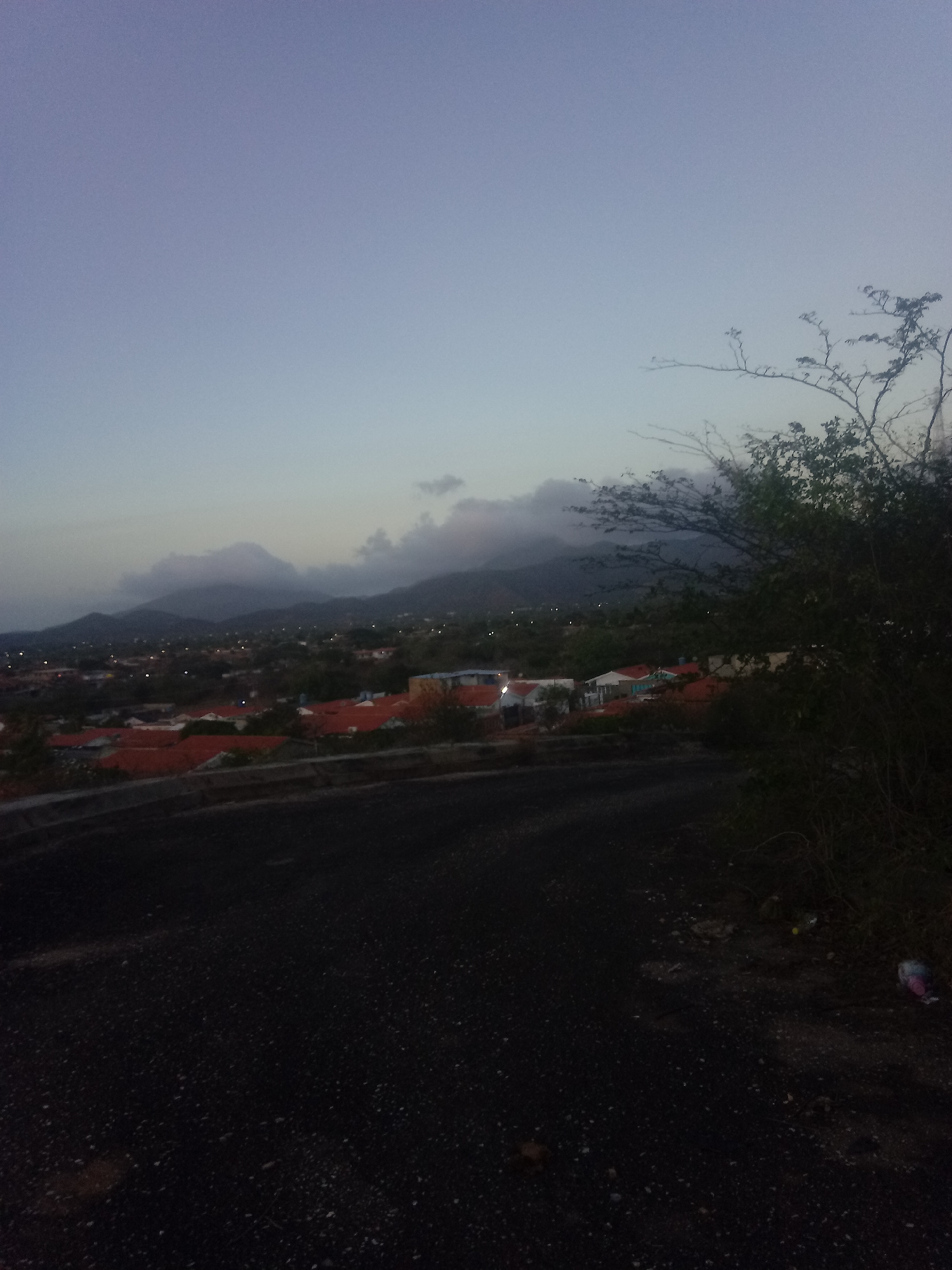 The image depicts a street view in Juan Griego, Venezuela, at dusk or dawn. Mountains are visible in the background, partially obscured by clouds. Below the mountains is a residential area with red-tiled roofs. In the foreground, a dark asphalt road is visible, with vegetation, possibly a tree or bushes, to the right. There are hints of litter scattered along the roadside. The sky is a gradient of light blue to grey.