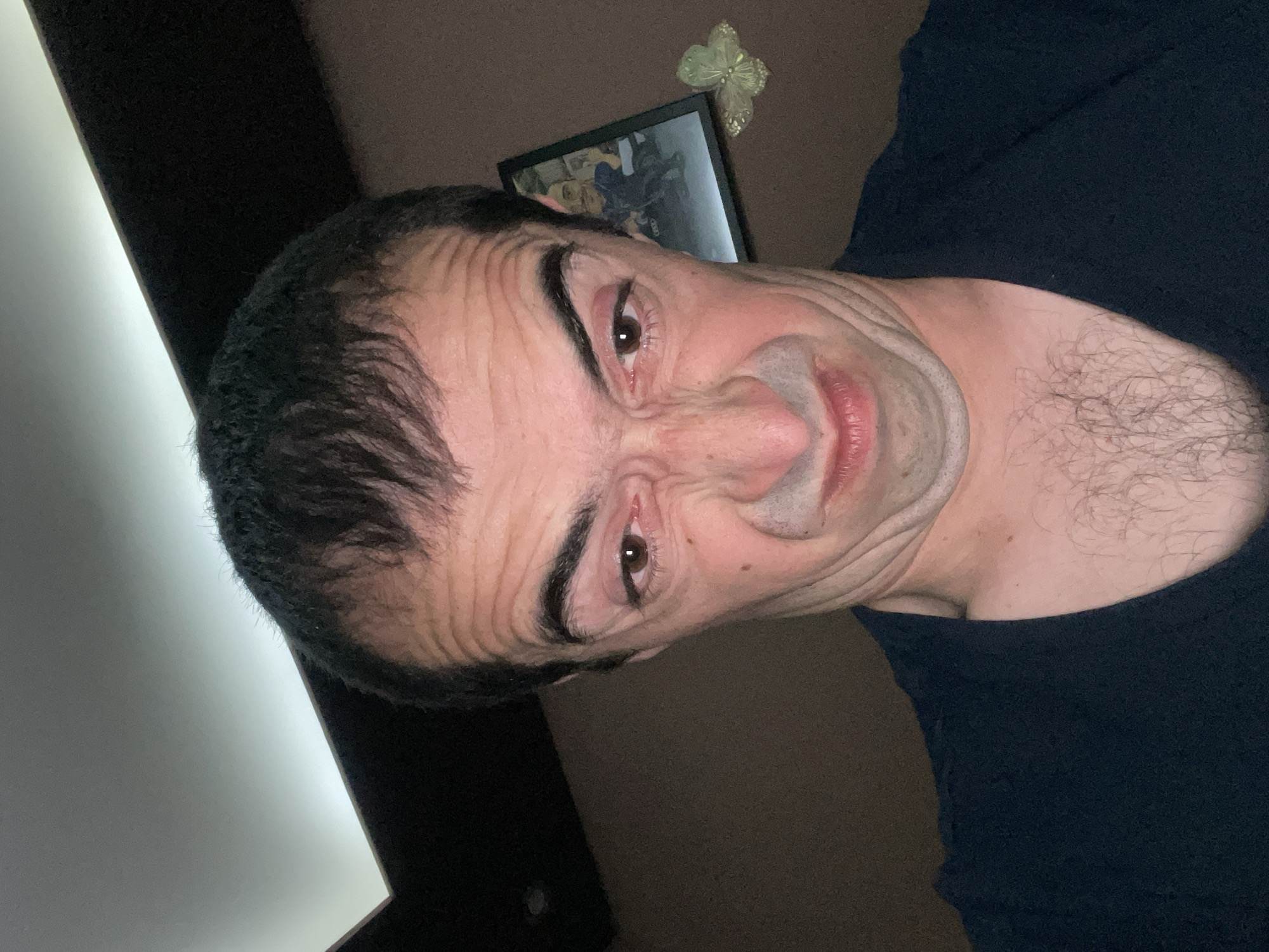 A man is making a face at the camera. He has brown eyes, dark hair, and a wrinkled forehead. He is wearing a dark shirt. In the background, there is a picture frame and a decorative butterfly on the wall. The location of the image is not specified and cannot be inferred from the image.
