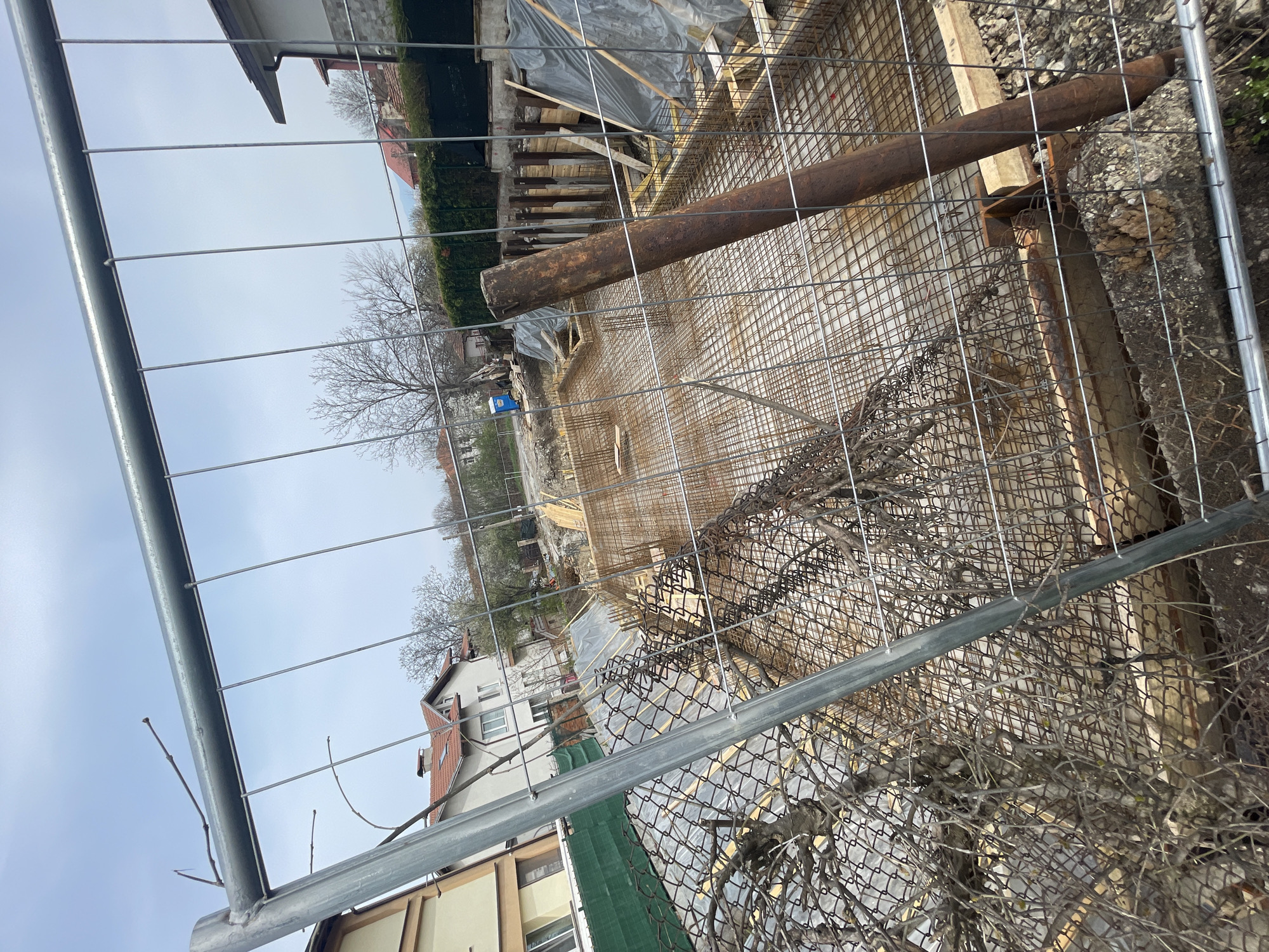 The image shows a construction site in Sofia, Bulgaria, viewed through a temporary chain-link fence. The site features a prepared foundation with rebar grid reinforcement, wooden formwork, and a large, rusty pipe placed across the structure. Visible beyond the construction site are residential buildings, trees with bare branches, and a blue portable toilet. The sky is overcast. The fence in the foreground is entwined with dead vegetation.