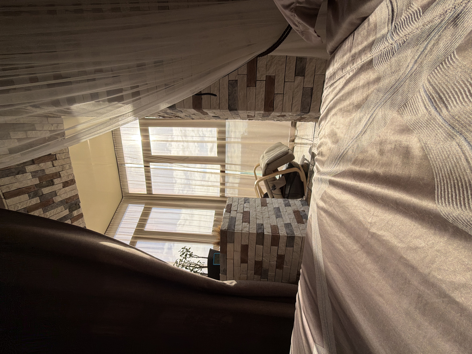 This image captures an interior scene with a prominent view of a balcony or sunroom through sheer curtains. The perspective is low, suggesting the photograph was taken from a bed. The foreground is dominated by a textured duvet or bedspread, with diagonal light and shadow patterns.
The middle ground features two stone-clad structures. The one on the left, closer to the viewer, is a rectangular column topped with a small potted plant. The one on the right is also a rectangular column. Between these structures, a light-colored rocking chair with a light-colored cushion is visible.
The background reveals a floor-to-ceiling window or series of windows, revealing a bright sky with clouds. The window frames are light-colored. The ceiling is also visible, with a textured surface that appears to be stone or tile. The overall ambiance is bright and airy, with the sunlight creating a warm glow.
The location is identified as Surgut, Russia. The presence of a bedroom, a sunroom area, and the specific architectural style might indicate a residential apartment or house in Surgut. There are no people visible in the image. The scene is static and appears to be a still life composition of a well-lit interior space.