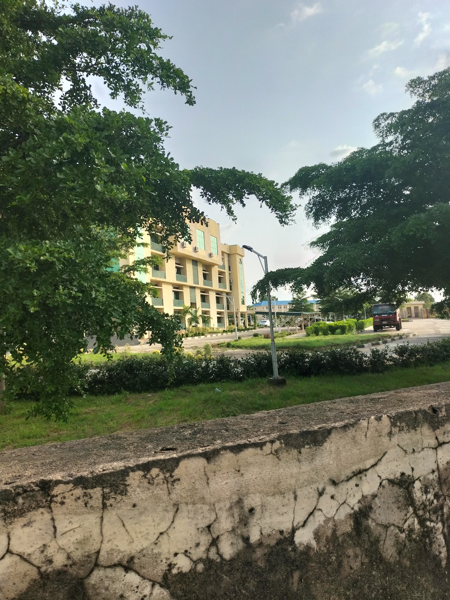 This is an eye-level, medium shot photograph taken outdoors in Jalingo, Nigeria. In the foreground, a cracked, weathered concrete wall runs horizontally across the bottom of the frame, textured with patches of dark moss and lighter, aged concrete. Behind the wall, a neatly manicured lawn transitions into a landscaped area with various bushes and low-lying greenery. A lamppost stands prominently in the middle ground, its dark pole topped with a light fixture.
To the left, a multi-story building with a beige facade and prominent green-tinted windows rises. The building appears to be modern, featuring several floors with visible balconies or protruding sections. Air conditioning units are visible on some of the windows. A lush, mature tree with abundant green foliage arches over the left side of the scene, partially obscuring the building.
In the background, beyond the landscaped area and the lamppost, a red Isuzu truck is parked on a paved surface. Further in the distance, additional low-lying buildings with blue roofs are visible, and the paved area suggests a road or parking lot. The sky above is partly cloudy, with a mix of blue sky and white clouds, indicating daylight conditions. The overall scene depicts an urban or institutional setting with greenery and architecture.