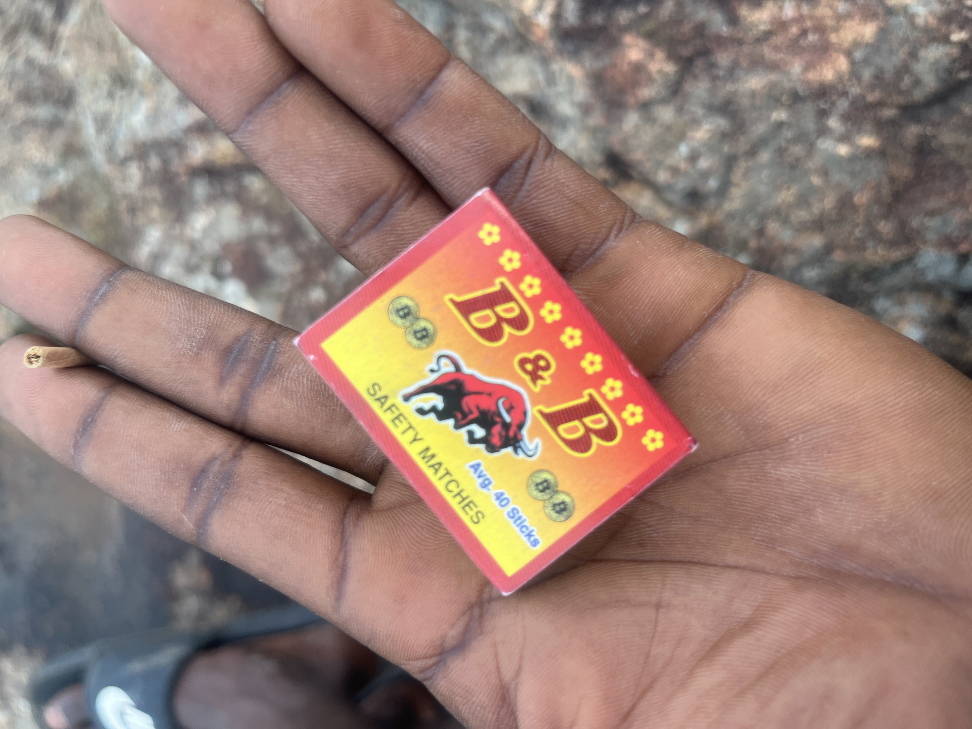 A person's hand, with dark skin and visible wrinkles, holds a small box of "B & B Safety Matches" in Gura Top, Nigeria. The matchbox is predominantly yellow and orange, featuring a red illustration of a devil-like figure and the text "SAFETY MATCHES" and "Avg. 40 Sticks". The background consists of rough, rocky terrain.