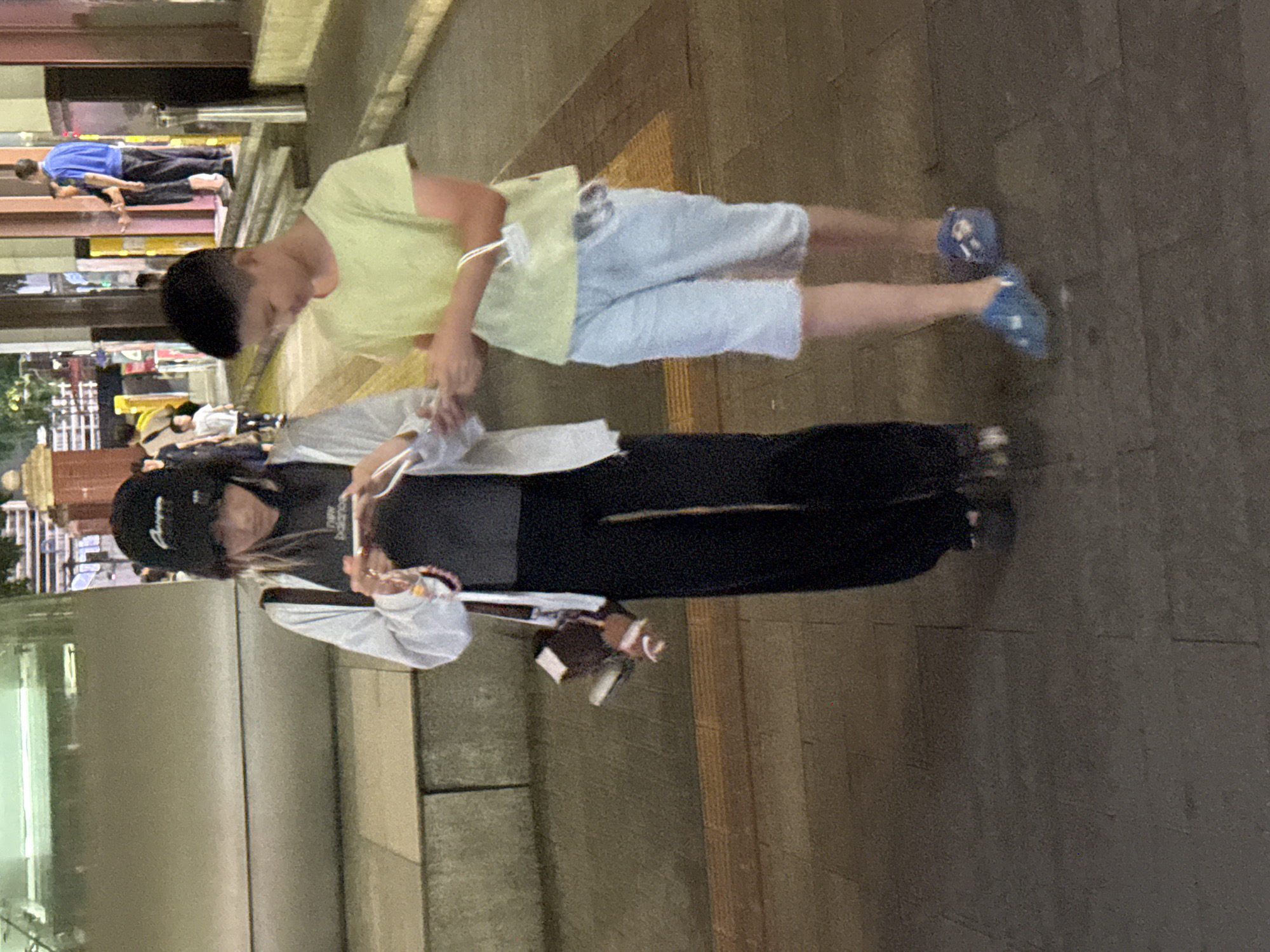 In the city of Yuzhong District, China, at street level, two young individuals, a female and a male, are walking. The female, on the left, wears a black baseball cap, a white jacket over a dark t-shirt, and black pants. She is looking down at a mobile phone in her hands. A dark handbag with a patterned strap is slung over her shoulder. The male, on the right, has short dark hair and is wearing a light yellow t-shirt and light blue knee-length shorts. He is also looking at something in his hands, possibly interacting with the female. Both are wearing casual footwear. The environment appears to be an outdoor urban setting, with textured paving stones on the ground and architectural elements, possibly a building facade, visible in the background. The lighting suggests it is evening or nighttime.