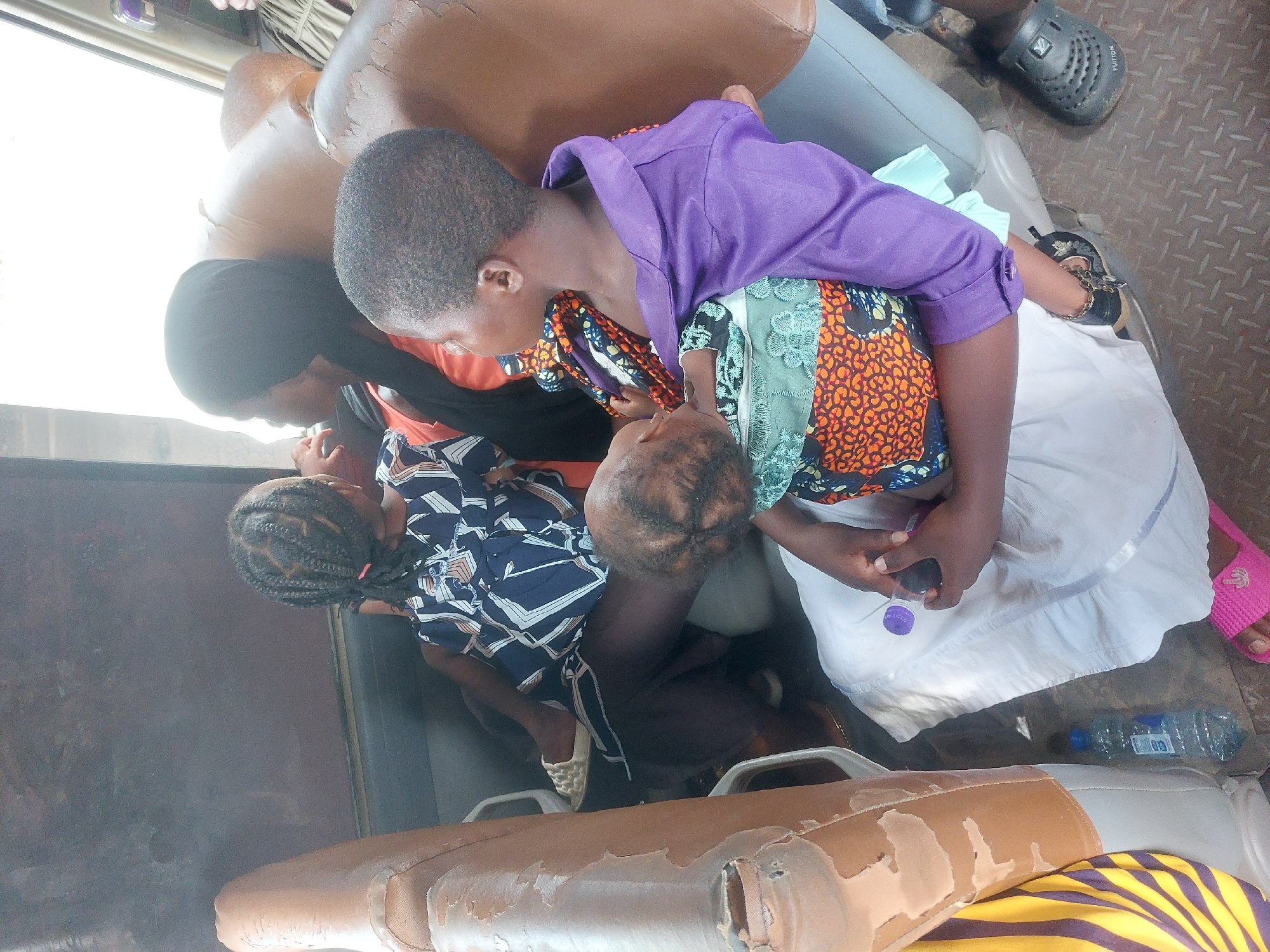 The image shows a group of four people inside what appears to be a vehicle, possibly a bus or a van. One woman wearing a black hijab is seated next to a child with braided hair. Next to them, a woman in a purple shirt and colorful patterned clothing is holding a baby or small child. A man with short hair is also present in the scene. A water bottle is visible on the floor. The specific location in Dar es-Salaam, Tanzania is not identifiable from the image.