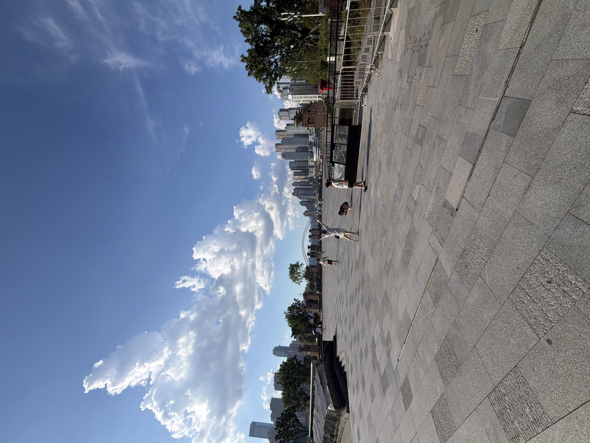 In Yuzhong District, China, a wide, sunlit plaza paved with large granite tiles stretches towards a cityscape dominated by towering modern buildings. A clear blue sky with scattered white clouds forms the backdrop. In the foreground and mid-ground of the plaza, several individuals are present. A young person in a white dress is raising their arms towards the sky. Nearby, another person is walking with a dog. Further back, more figures are scattered across the plaza, some appearing to walk and others in conversation. Trees line the edge of the plaza, and a metal railing with stairs is visible on the right side of the image. In the distance, a modern bridge spans across a body of water, connecting to the urban skyline. The overall scene depicts a public space in a bustling city, bathed in daylight.