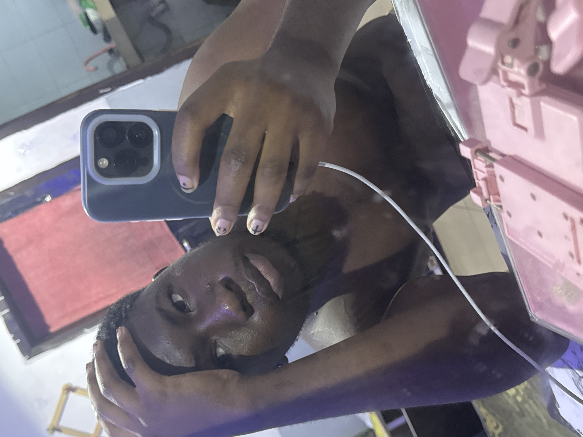A person is taking a selfie in a mirror. The person is dark-skinned and has short, dark hair. They are holding a phone with a gray case and three camera lenses. There is a white cord visible near the person's neck. A pink object with hinges is visible on the right side of the mirror. The reflection shows a room with blue and red elements, possibly curtains or decor. Parts of the floor and other objects are also visible in the mirror reflection, but they are not clear. There is no visible information indicating that the image was taken in Ipetumodu, Nigeria.