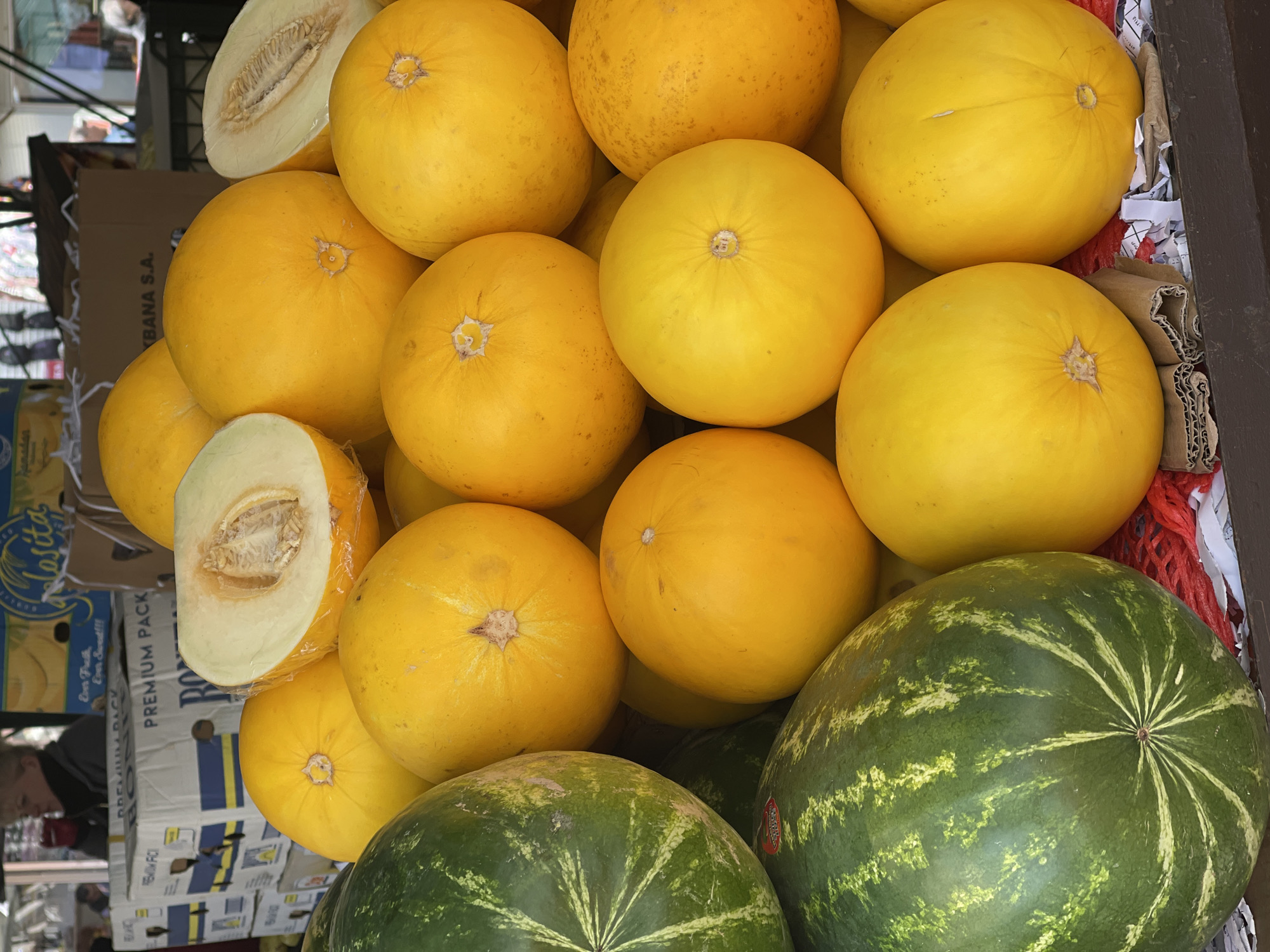 The image shows a pile of yellow cantaloupes and green watermelons in a market stall. The cantaloupes are mostly whole, but a couple of them are cut in half, revealing the inner flesh and seeds. The watermelons are clustered at the bottom of the pile. The melons are in a cardboard container, part of which is visible on the left, with the writing "PREMIUM PACK" and "BANA S.A." visible on it. There are white papers and red netlike structures among the fruits. The setting appears to be a street market.