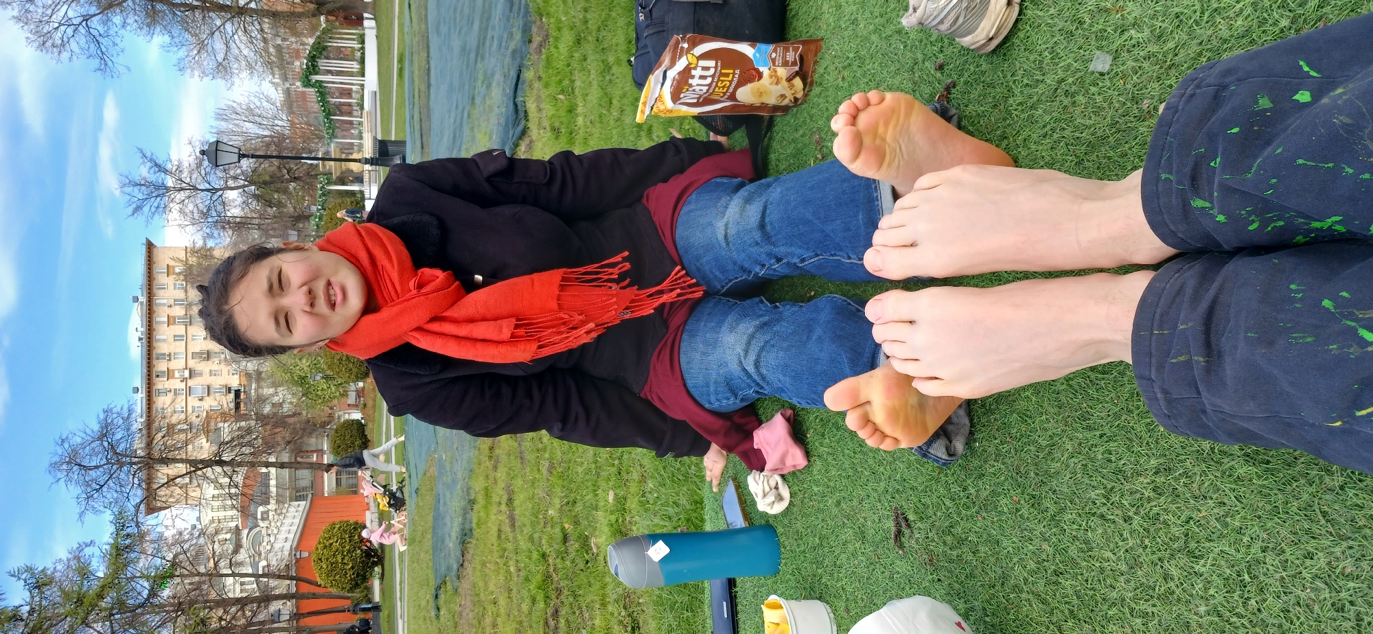 A medium shot depicts two people reclining on a grassy area, their bare feet prominently displayed. One individual, a young woman, is seated and facing the camera, wearing jeans, a black jacket, and a bright orange scarf. Her eyes are partially closed, and she has a slight smile. Beside her sits another person, clad in dark pants splattered with green paint, with only their bare feet visible. A bag of "Natti Vesli" granola is lying near the feet.
The backdrop includes an urban park setting, with buildings visible in the distance. Notably, the scene is situated in Moscow, Russia, recognizable by the architecture of the buildings and the specific design of the street lamps. The sky is clear with sparse clouds, suggesting a bright day. In the immediate foreground, various personal belongings such as water bottles and clothing items are scattered on the ground.