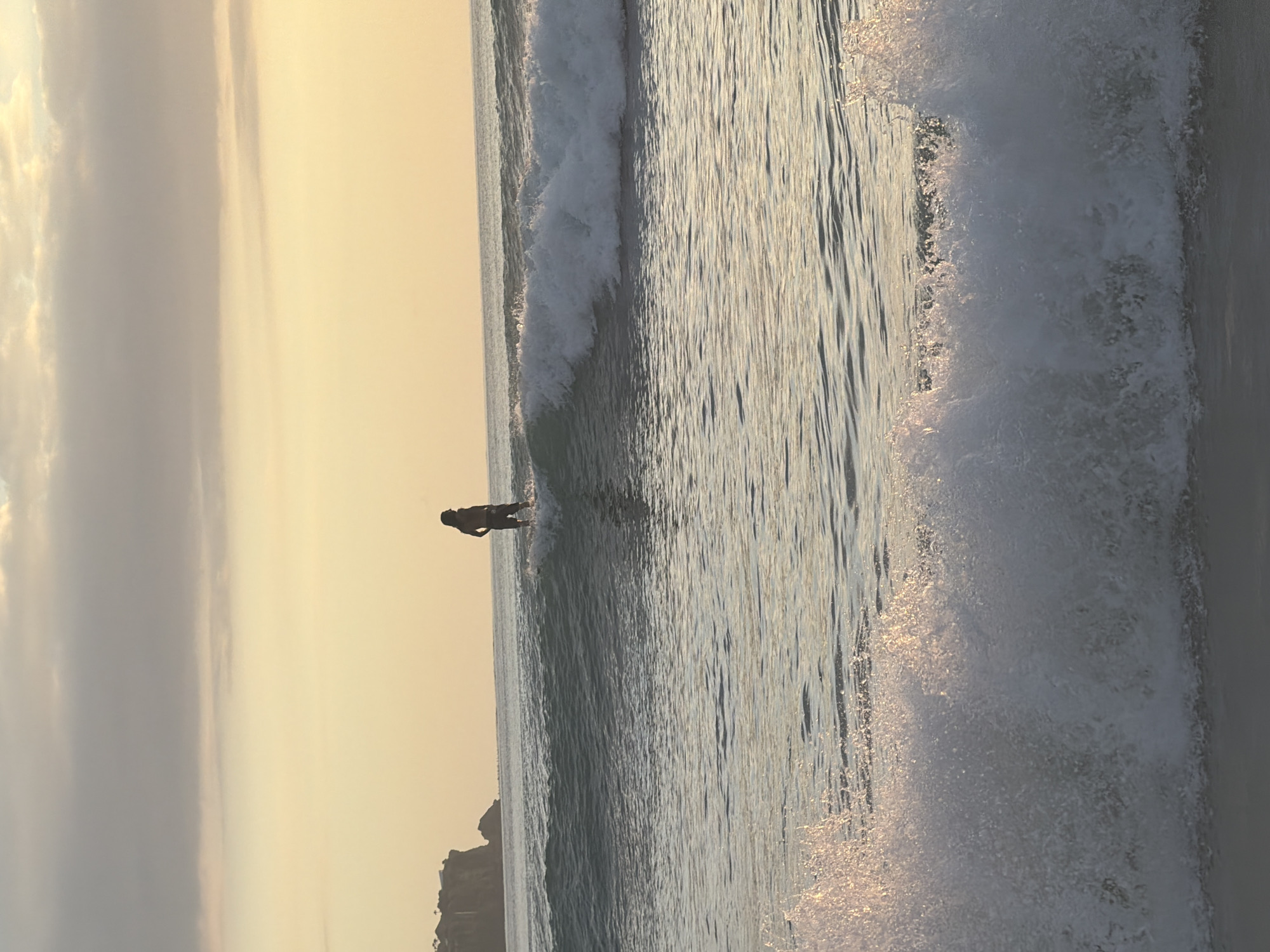 A person is surfing in the ocean at sunset in Kuta Selatan, Indonesia. The sky is a hazy yellow-orange, and there are clouds in the distance. The ocean is a mix of blue and white as the waves break. A rocky outcrop is visible in the distance. The person is silhouetted against the bright sky.