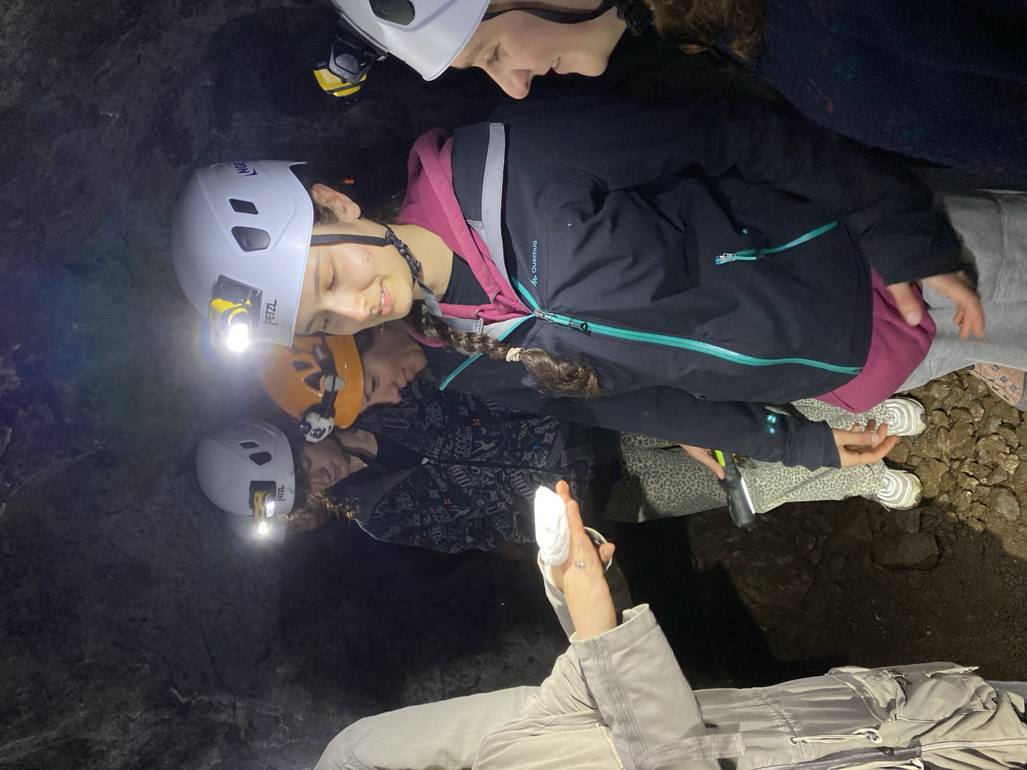 This image depicts a group of individuals, primarily young people (children or teenagers), engaged in an underground exploration activity, likely caving, in a dark, subterranean environment. The scene is illuminated almost exclusively by the headlamps worn by the participants, casting strong highlights and deep shadows.
The subjects wear protective gear typical for caving, including white and orange helmets. Several headlamps are visible, with one white helmet clearly showing the "PETZL" brand. Their attire consists of practical outdoor clothing, such as a black jacket with a prominent turquoise zipper and "QUECHUA" branding, and patterned grey trousers.
In the foreground, an adult's hands are visible holding a small, irregular white object, possibly a rock sample or mineral, which appears to be the focal point of the group's attention. Another person's hand holds a small, black flashlight. The ground beneath them is uneven, composed of dark, rocky terrain and dirt. The expressions on the visible faces are generally engaged and curious, with some individuals smiling, suggesting an enjoyable and educational experience.
The overall setting is one of complete darkness outside the immediate reach of the headlamps, indicating a cave or mine environment. The event taking place is an exploration or discovery activity, focused on observing the geological features or specimens found within the cave. The location context of Les Chavannes-en-Maurienne, France, aligns with such outdoor and underground activities.