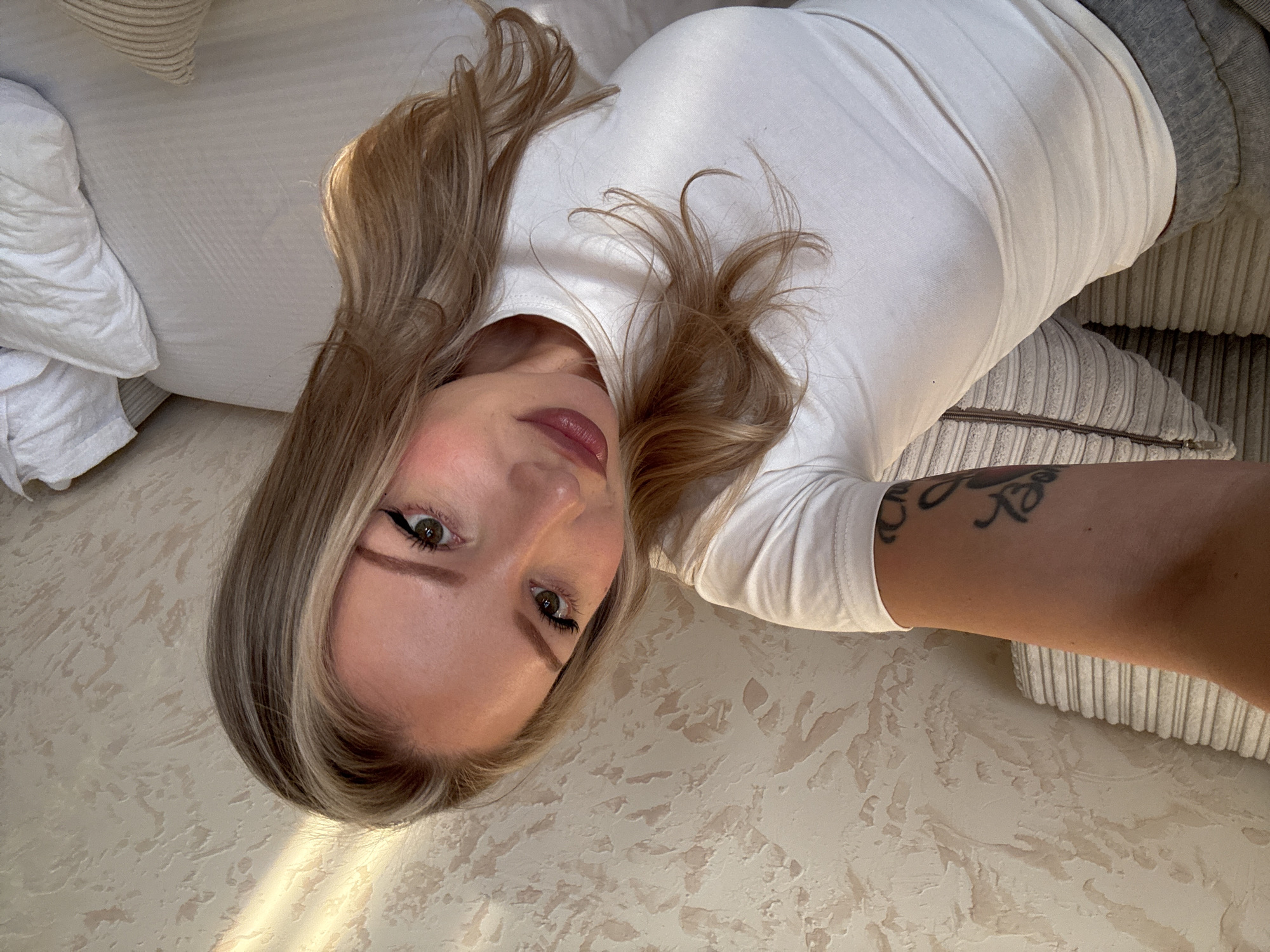 An overhead shot captures a woman lying on her back, looking directly towards the camera in an indoor setting in Baksan, Russia. She has long, wavy blonde and light brown hair. She is dressed in a white t-shirt and grey bottoms. Her facial features are highlighted with winged eyeliner, mascara, defined eyebrows, and pink/mauve lipstick, complementing her green/hazel eyes. A black script tattoo is visible on her right forearm. She is positioned on a light-colored surface, appearing to be a bed or sofa, with white bedding or fabric and a grey ribbed cushion. The background consists of a light beige, textured wall, possibly stucco or plaster. A patch of natural sunlight falls across her hair and the wall, indicating it is daytime. The overall scene suggests a relaxed and casual moment indoors.