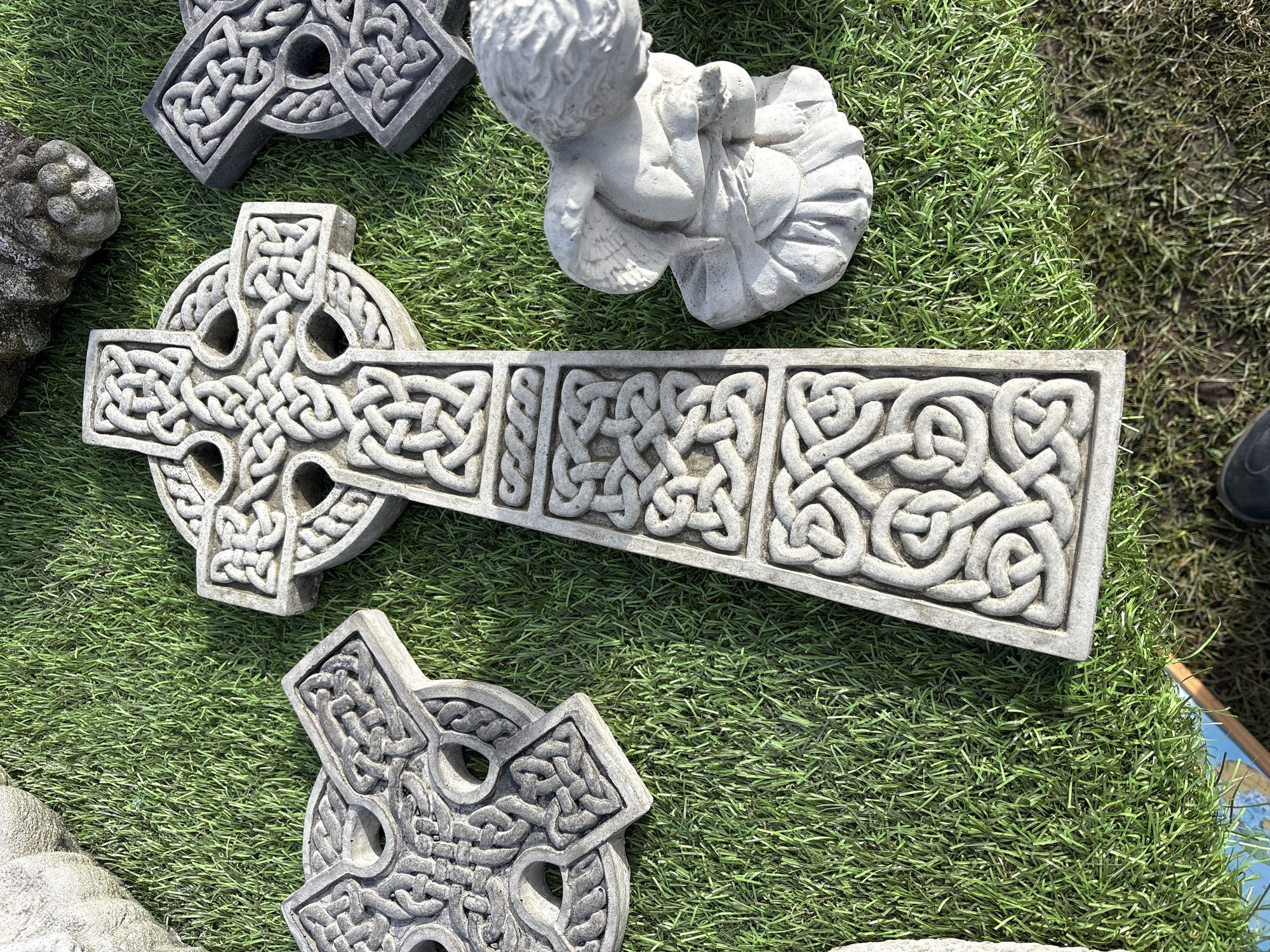 The image shows a collection of stone objects resting on artificial green turf. Prominently featured are three Celtic crosses, each with intricate knotwork designs. A small, weathered statue of an angel is also present. The objects appear to be displayed for sale, possibly in an outdoor setting in Chirk, United Kingdom. The lighting suggests a sunny day, with visible shadows cast by the objects. The composition is a high-angle shot, giving a clear view of the items' details and placement.