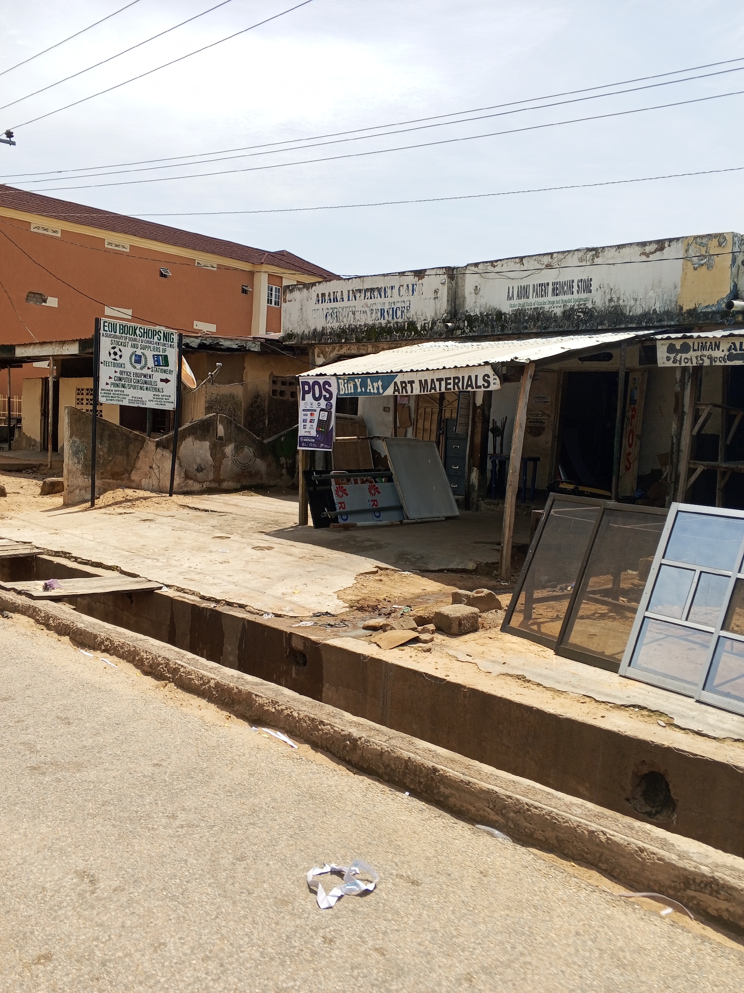 The image depicts a street scene in Jalingo, Nigeria, with several storefronts visible. On the left, a sign for "EOU BOOKSHOPS NIG" indicates a business selling textbooks, office equipment, computer consumables, and sporting materials. Adjacent to it, a building with a tiled roof and orange-colored walls is partially visible. In the center, a corrugated metal roof covers a series of stalls. One stall has a sign reading "POS" and "Bin Y. Art ART MATERIALS," suggesting services related to point-of-sale transactions and art supplies. Another storefront has a sign that is partially obscured but appears to read "LIMAN, AL". In front of these stalls, several large window frames, possibly for sale, are leaning against a concrete barrier. A drainage channel runs along the edge of the street in the foreground, with some debris present. Overhead, power lines crisscross the sky. The overall environment appears to be an outdoor commercial area with a mix of older and newer structures.