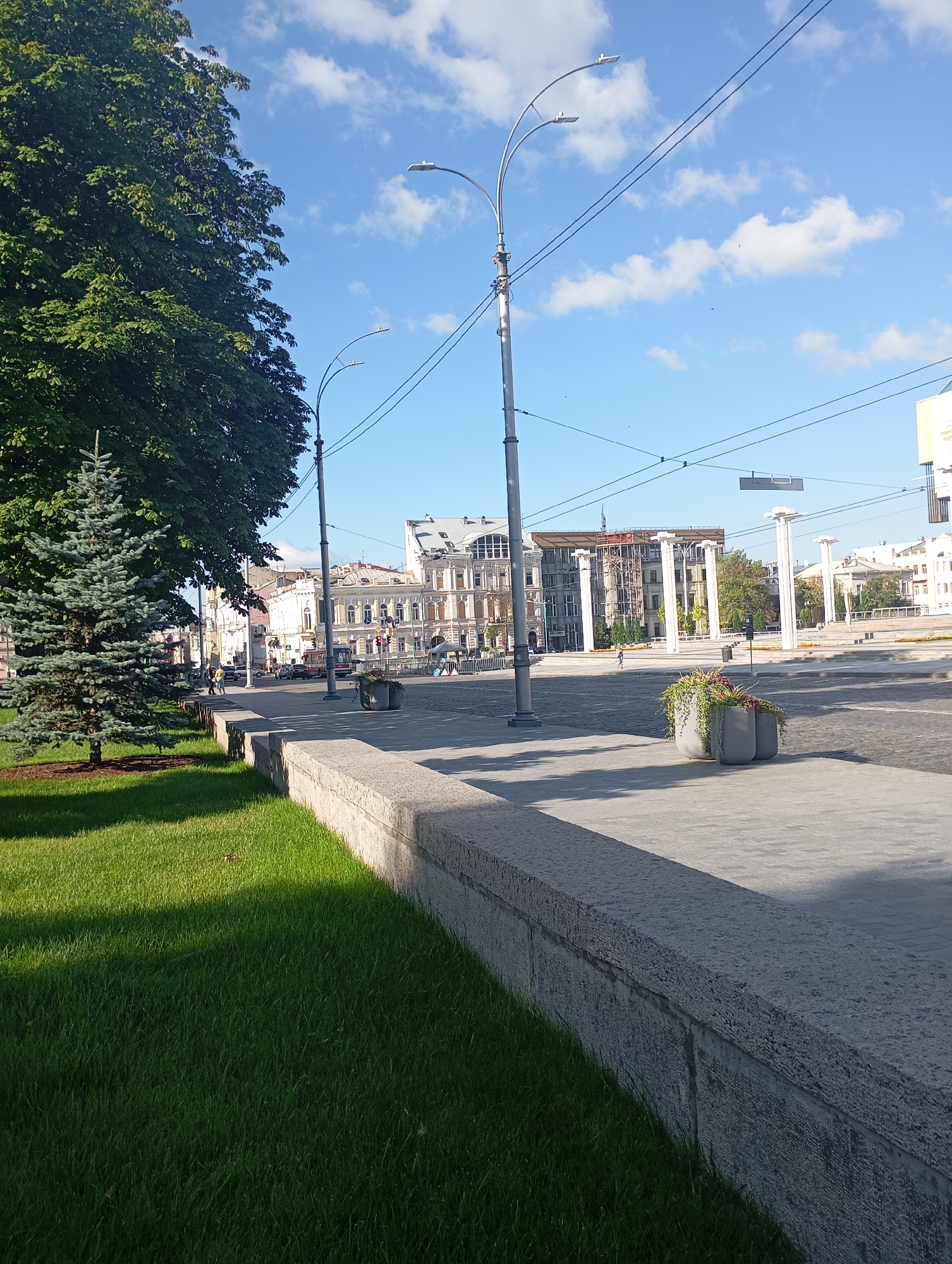 The image depicts a street scene in Kharkiv, Ukraine, specifically on Constitution Square. The foreground features a well-maintained grassy area with a young blue spruce tree. A low, textured stone wall runs along the edge of the grass, bordering a paved pedestrian walkway. Beyond the walkway, a wide street is visible with traffic lights, a trolleybus, cars, and several individuals walking.
In the mid-ground, classical-style buildings with ornate facades line the street. In the distance, a construction site is evident with scaffolding on one of the buildings. The sky is clear blue with scattered white clouds, and streetlights with multiple lamps are positioned along the thoroughfare. Two large grey planters filled with greenery are situated on the pedestrian area. The overall impression is of a bright, sunny day in an urban setting.