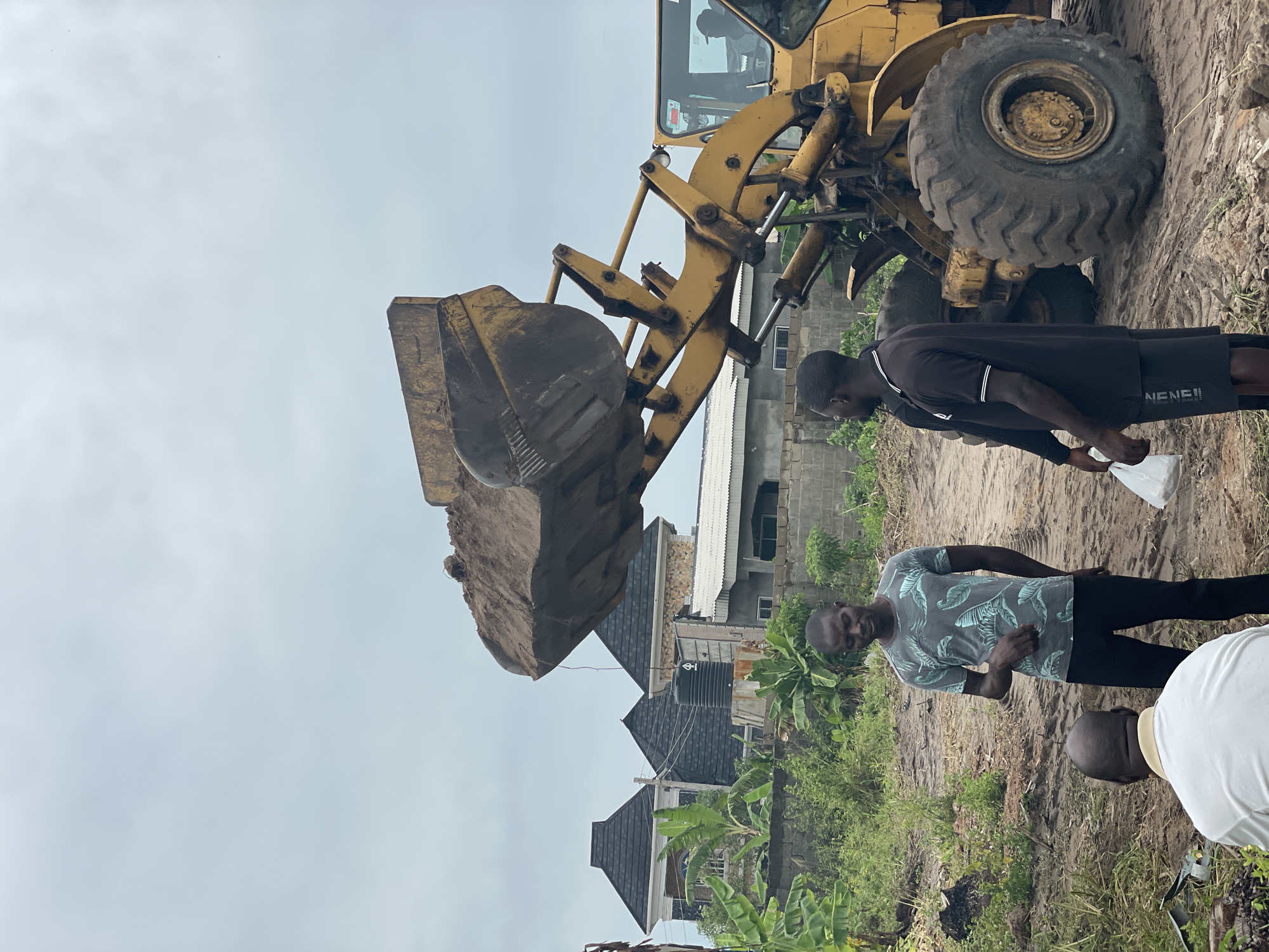 The scene depicts a construction site in Badagry, Nigeria. A yellow front-end loader is elevated with a full bucket of dirt. Two men are standing nearby. One man is wearing a black shirt and black shorts. Another man is wearing a gray shirt with a black pattern and black pants. A third man is partially visible, wearing a white shirt. Behind them are several residential buildings. The ground is dirt and vegetation. The sky is overcast.
