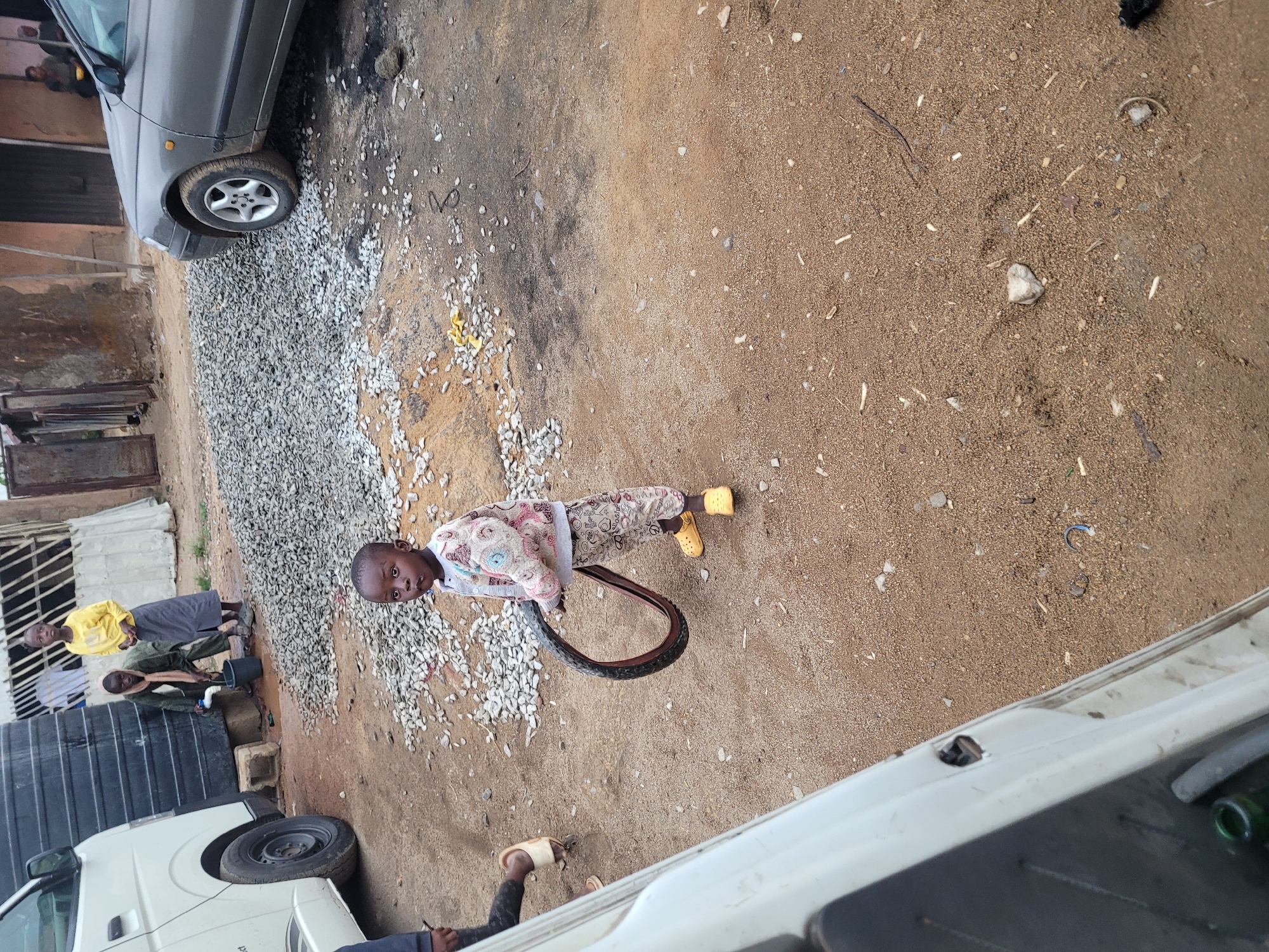 A child in a patterned shirt and yellow shoes holds a tire on a dirt and gravel ground in Jos, Nigeria. Other people are visible in the background near a white truck and a grey car.