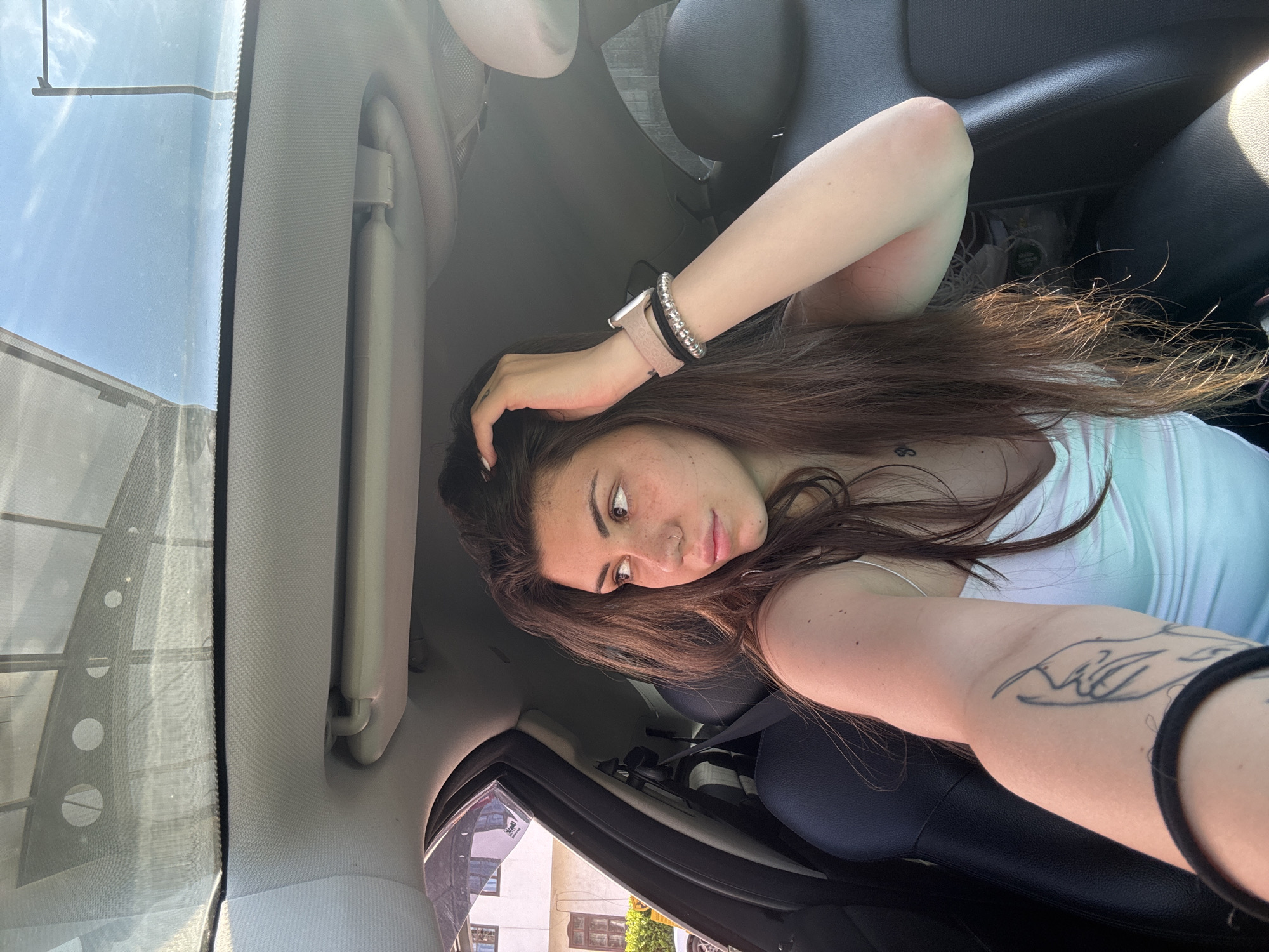 A young woman with long, brown hair and freckles is sitting in the passenger seat of a car, looking out the window. She is wearing a white tank top and has a tattoo on her left arm. She is also wearing a watch and bracelets on her right wrist. The car's sunroof is open, and the sky is visible. The scene appears to be in a city, possibly Sofia, Bulgaria, given the architecture visible outside the car window. The lighting suggests it is daytime, with sunlight streaming into the car.