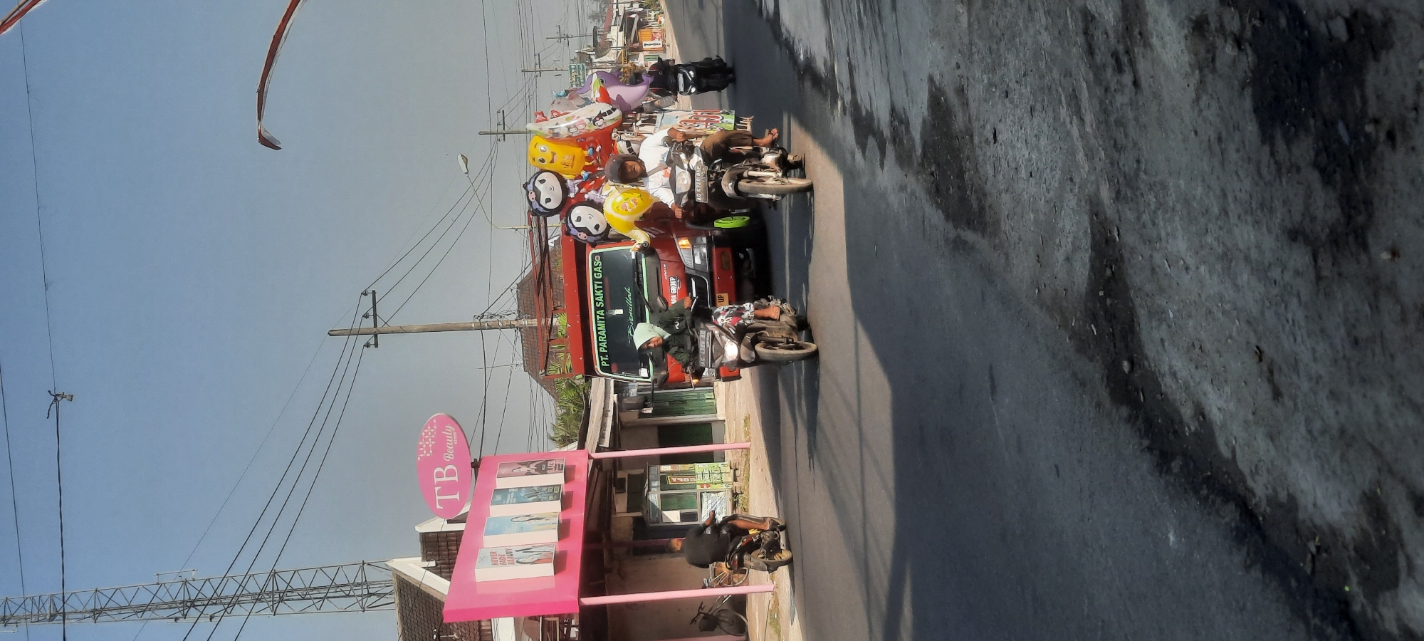 The image depicts a street scene in Selopuro, Indonesia, likely during daylight hours, given the clear sky and shadows. A red truck with the text "PT. PARAMITA SAKTI GAS" is prominently featured, carrying a load of colorful balloons. Several people are riding motorcycles. In the foreground, a person on a motorcycle with license plate "AG 6720 BBP" is heading towards the viewer. To the left, another person on a motorcycle is moving away from the viewer. Further to the right, a third motorcyclist is also moving away from the viewer. On the left side of the street, a pink building with the signage "TB Beauty" is visible, displaying several images on its facade. A tall communication tower stands in the background. The road surface appears to be asphalt with some patches of wear and tear. Electrical wires are strung across the scene, supported by utility poles. The overall impression is of a typical street in a residential or commercial area of a town in Indonesia.